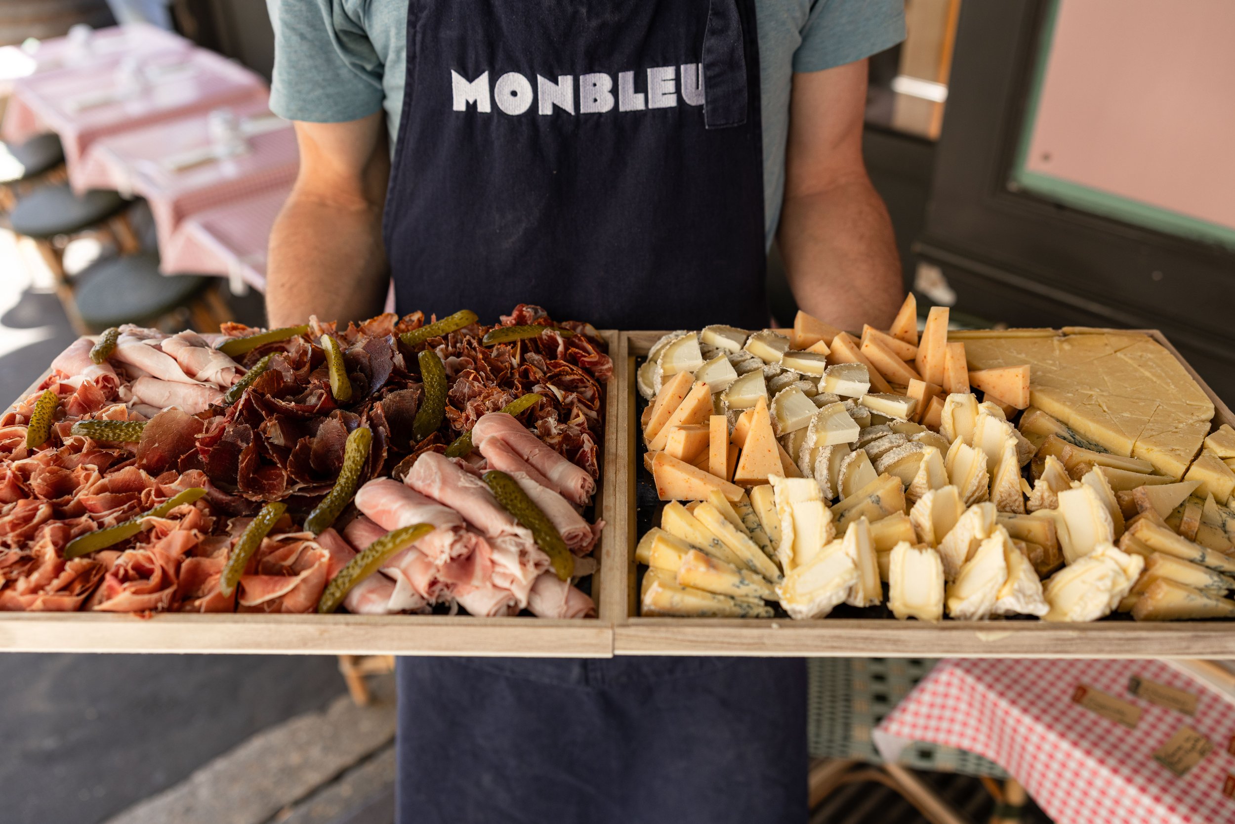 Un homme vêtu d'un tablier monbleu tient dans ses mains deux plateaux, un à gauche de charcuterie et un à droite de plusieurs sortes de fromage