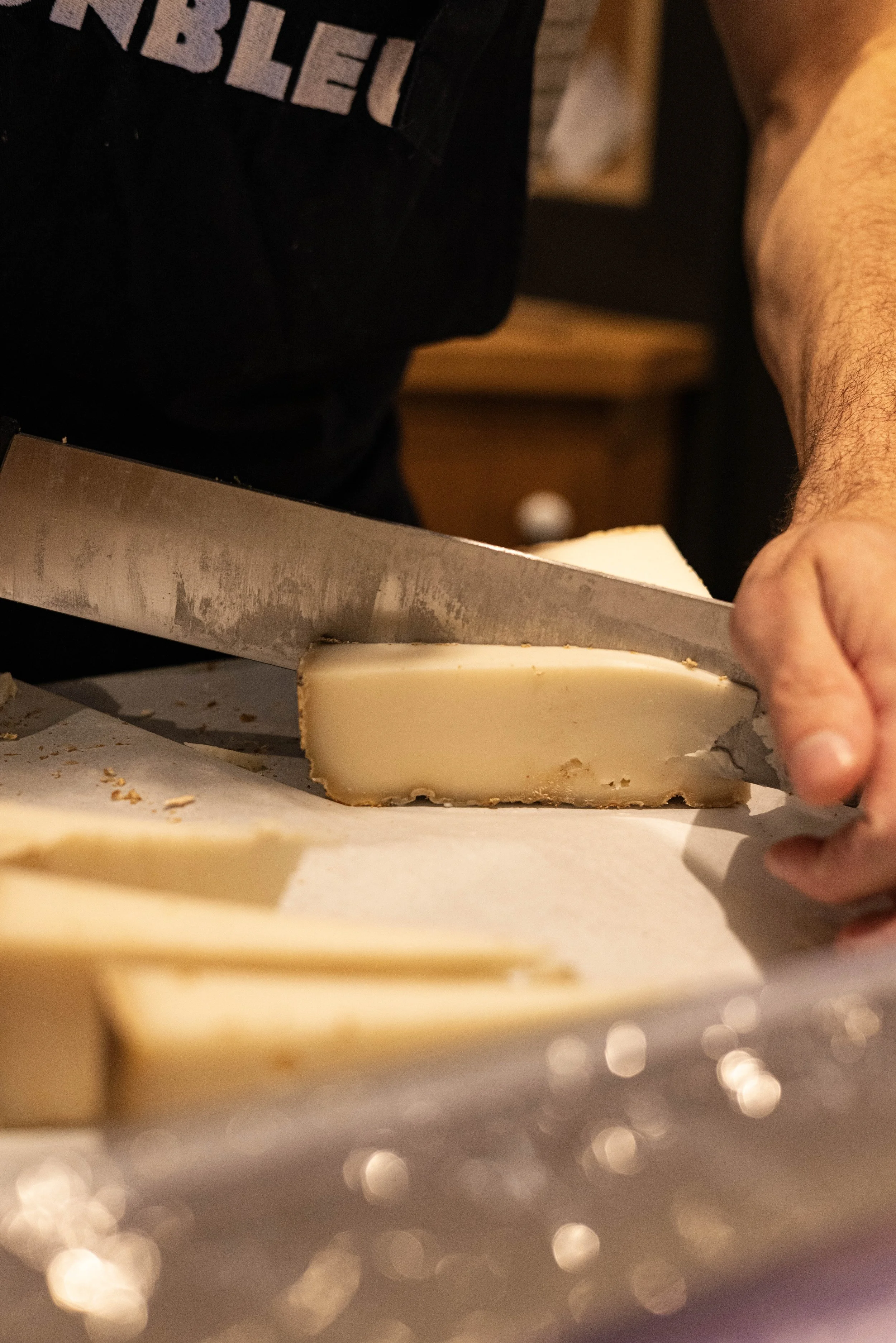 Personne coupant un morceau de fromage avec un grand couteau, dans un environnement intérieur.