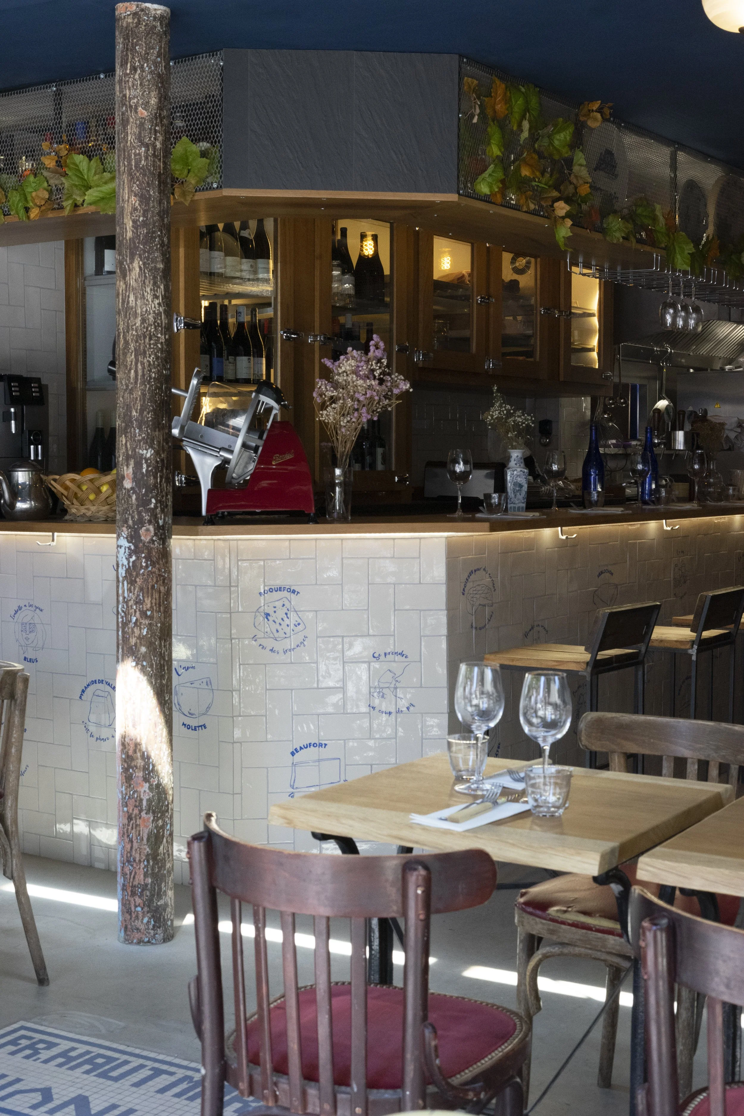 Intérieur d'un restaurant avec des tables en bois, verres, assiettes, et chaises en bois. Un comptoir de bar avec des bouteilles de vin, des fleurs décoratives, et une ambiance chaleureuse.