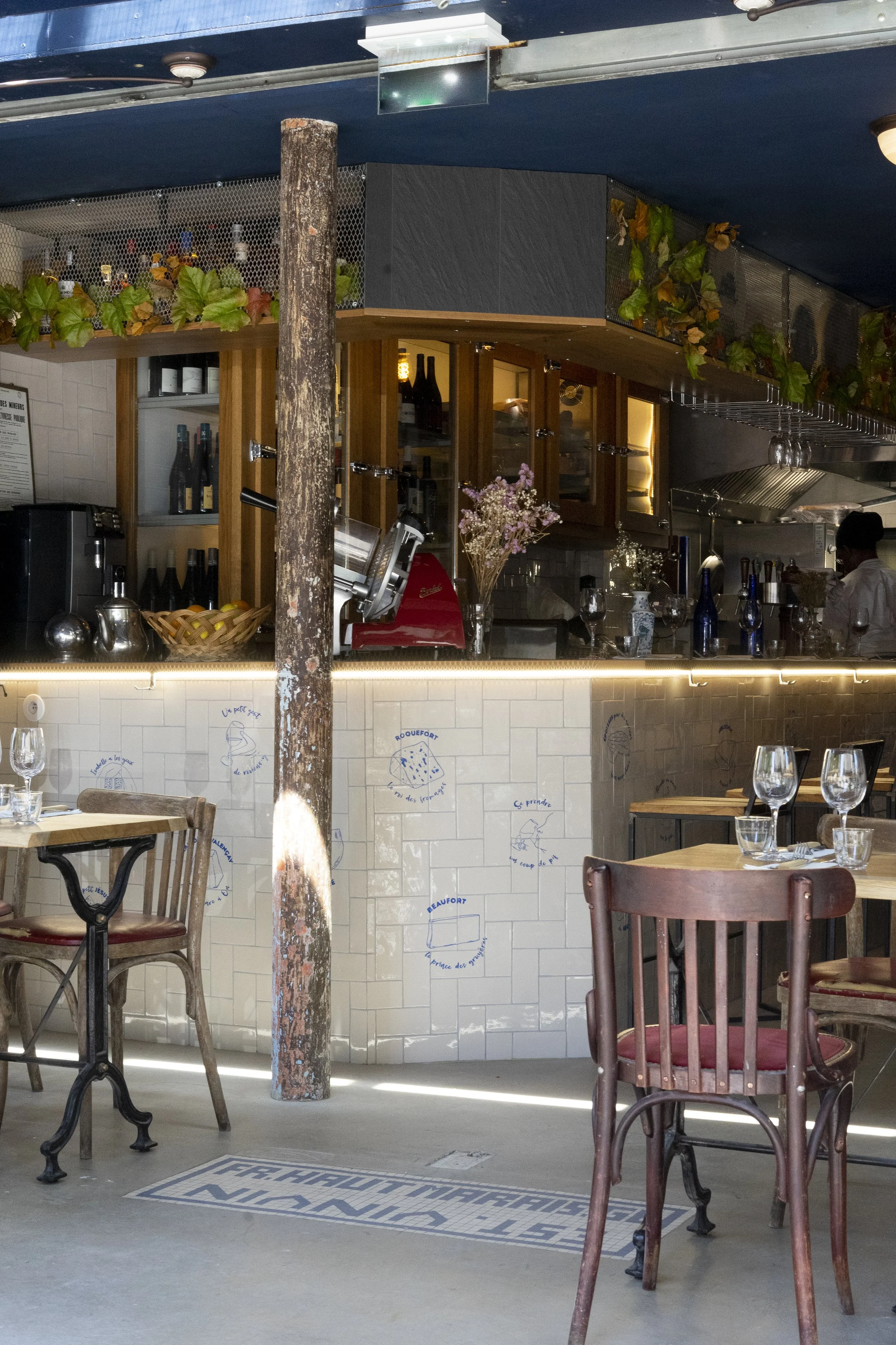 Intérieur d'un restaurant, un tronc d'arbre est devant le comptoir, les tables sont dressées, il y a des fleurs de disposées, on voit des bouteilles de vins, le restaurant est vintage