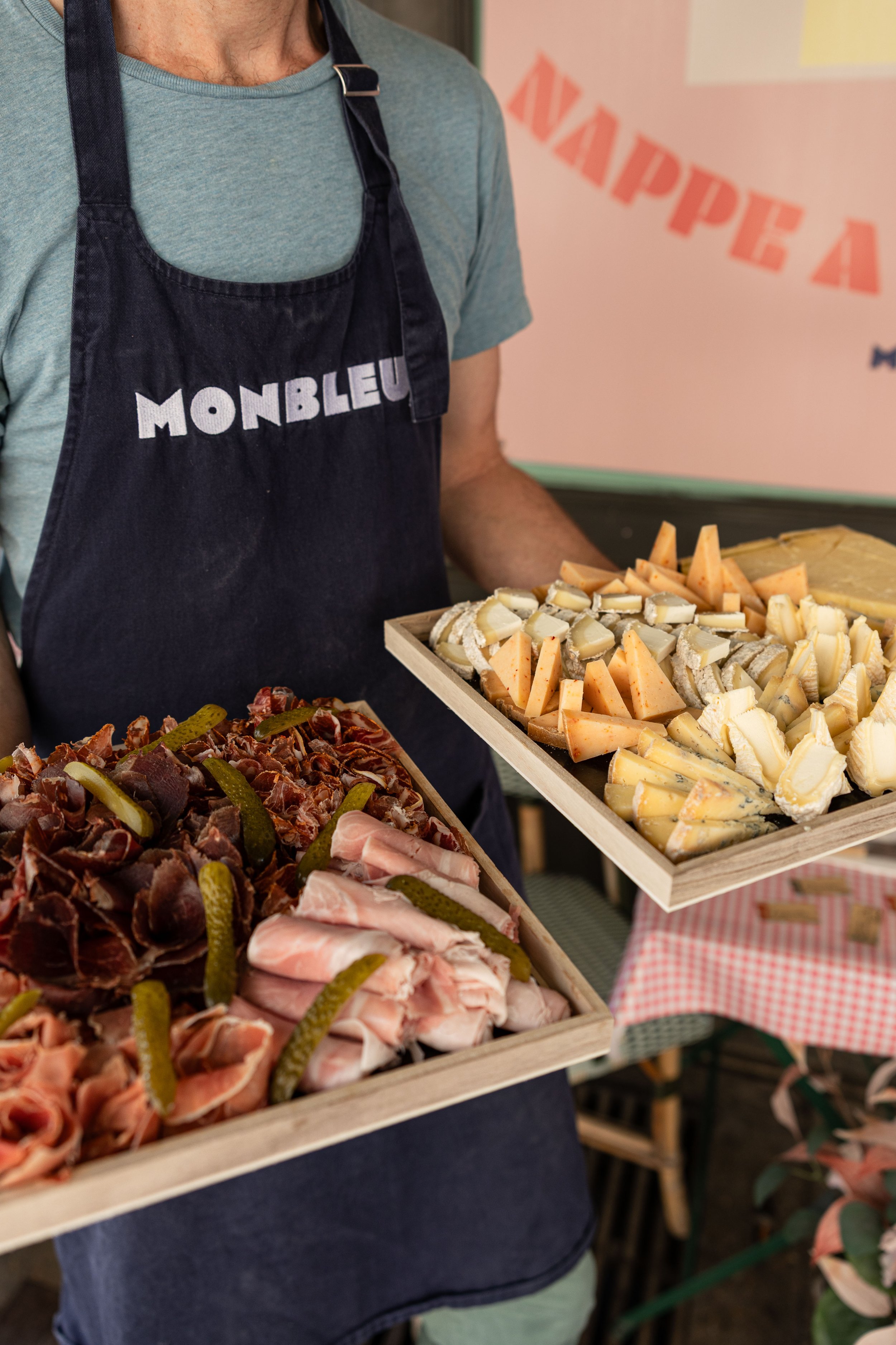 Un monsieur vêtu dans tablier "monbleu" tient deux plateaux, un rempli de plusieurs fromages et un autre, rempli de plusieurs charcuterie et des cornichons