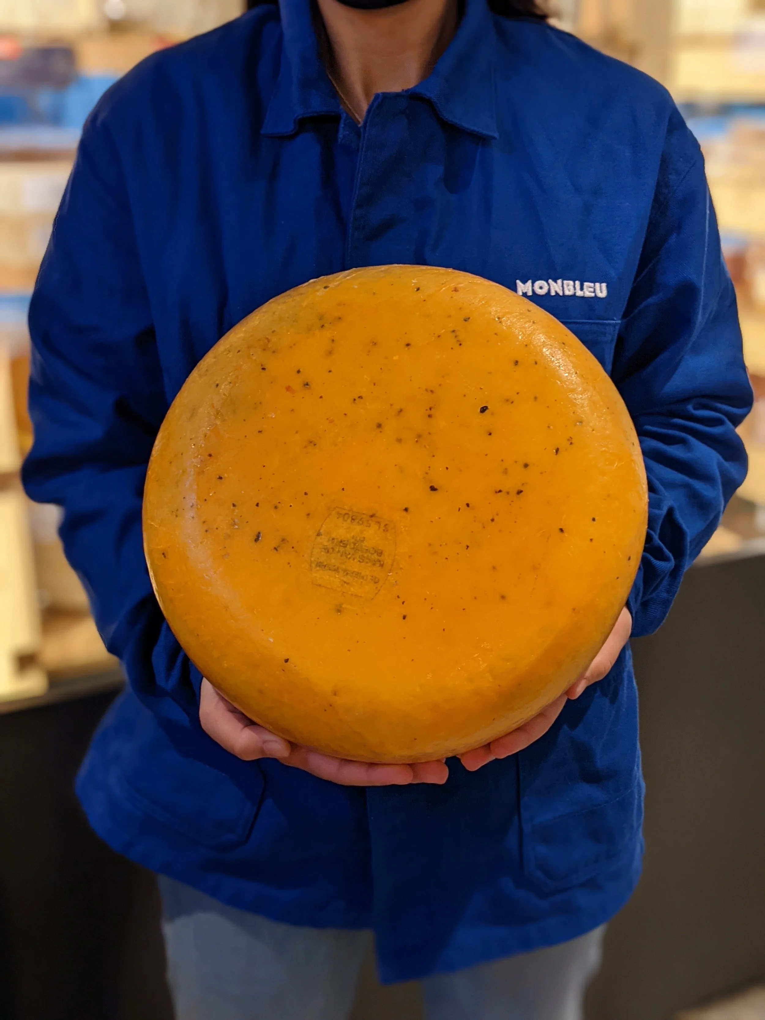 Une grosse meule de fromage est tenu par une personne portant une veste bleu électrique avec écrit Monbleu