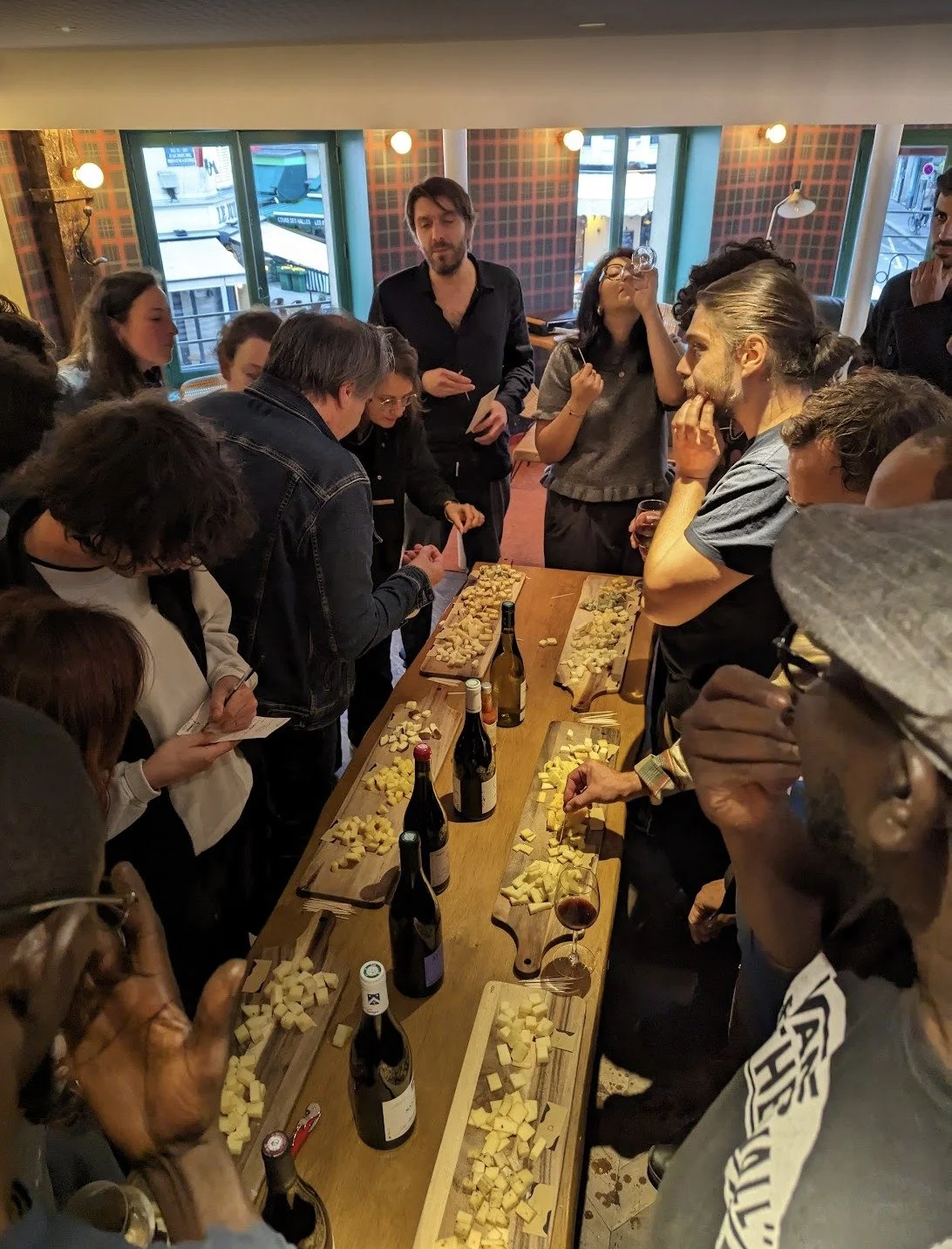 Grand buffet avec plusieurs planches à fromage ainsi que des bouteille de vins, beaucoup de personnes mangent debout autour de cette table et discutent