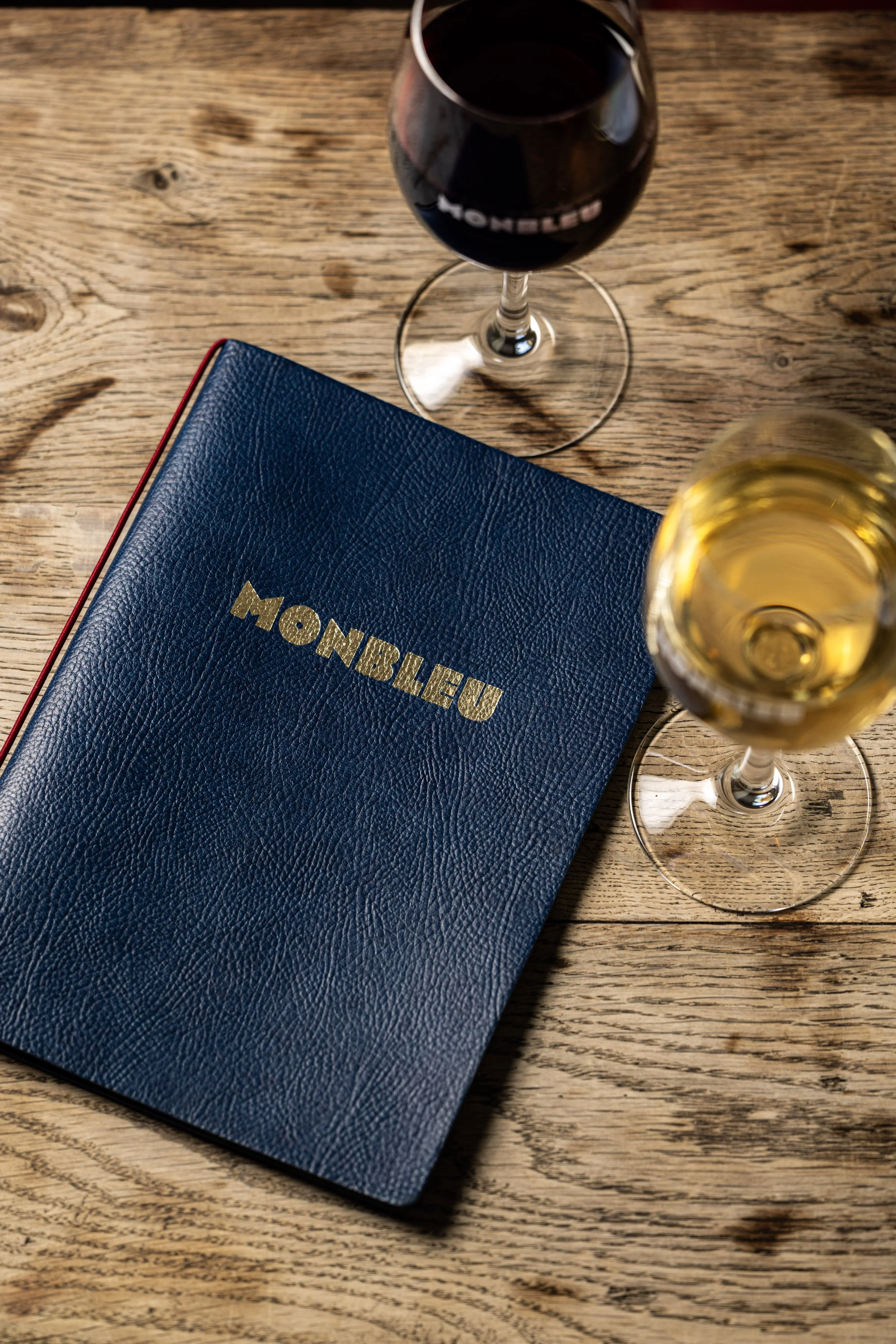 Un menu monbleu posé sur un table en bois et deux verres de vin, un de vin rouge et un de vin blanc 