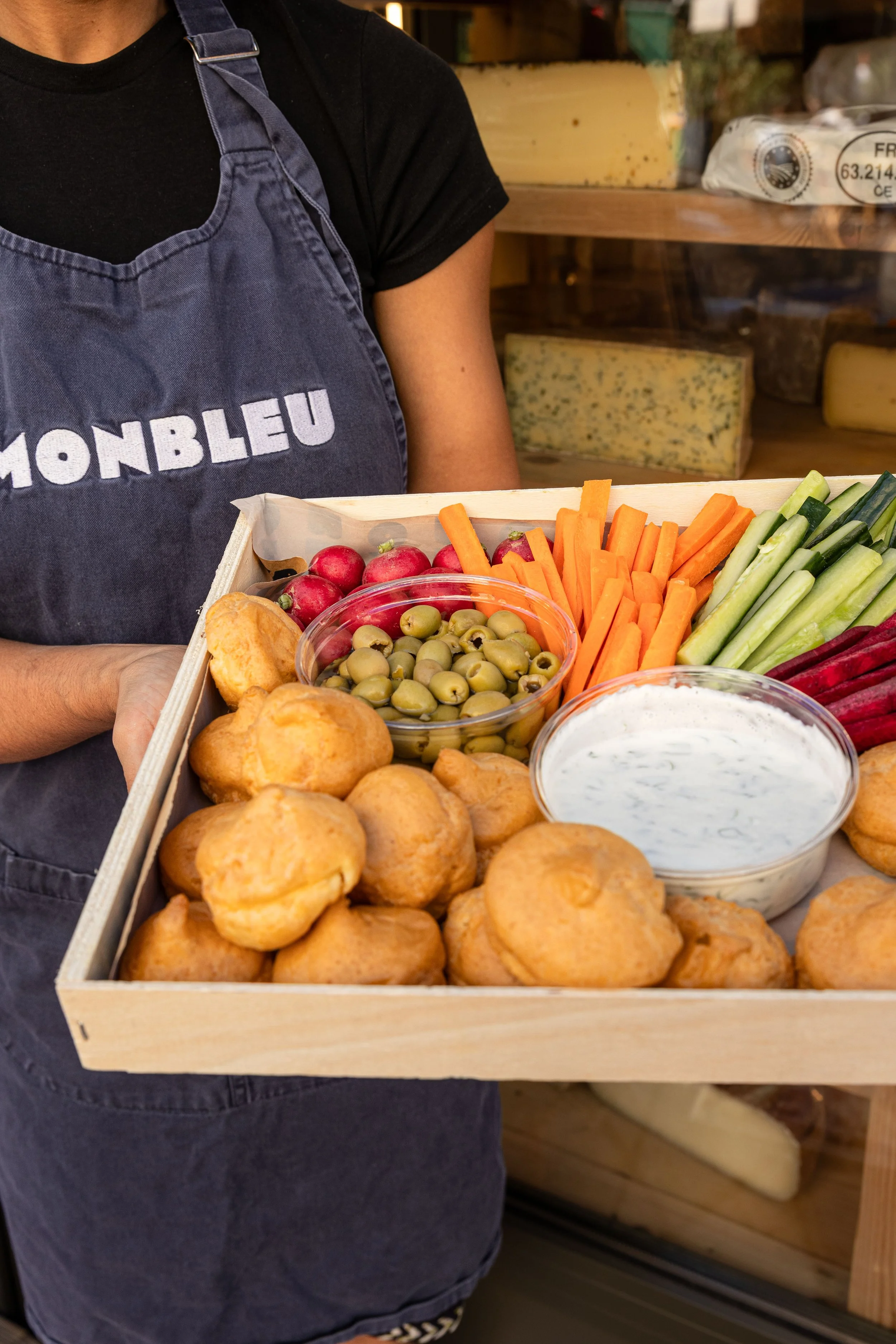 Une femme tient un plateau rempli de nourriture pour un apéro, gougères, olives, sauce blanche, carottes, radis et concombres