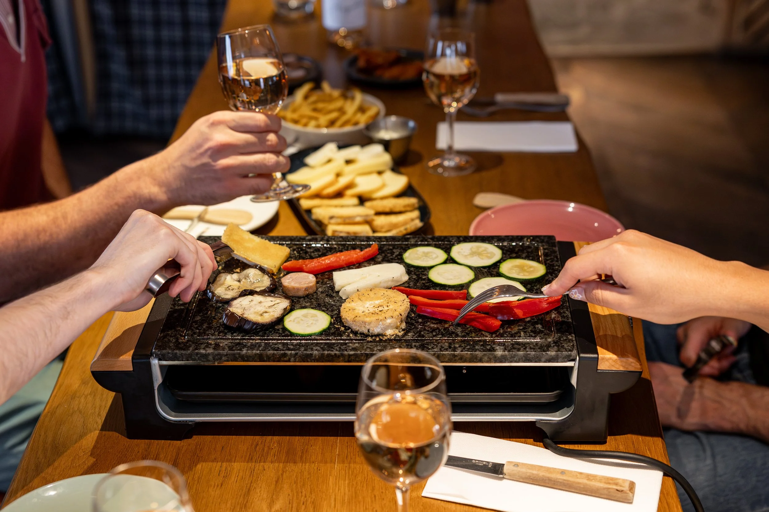 Plusieurs personne autour d'une table partage un repas, il font griller leurs aliments sur une plancha, il y a des verres à vin et des assiettes