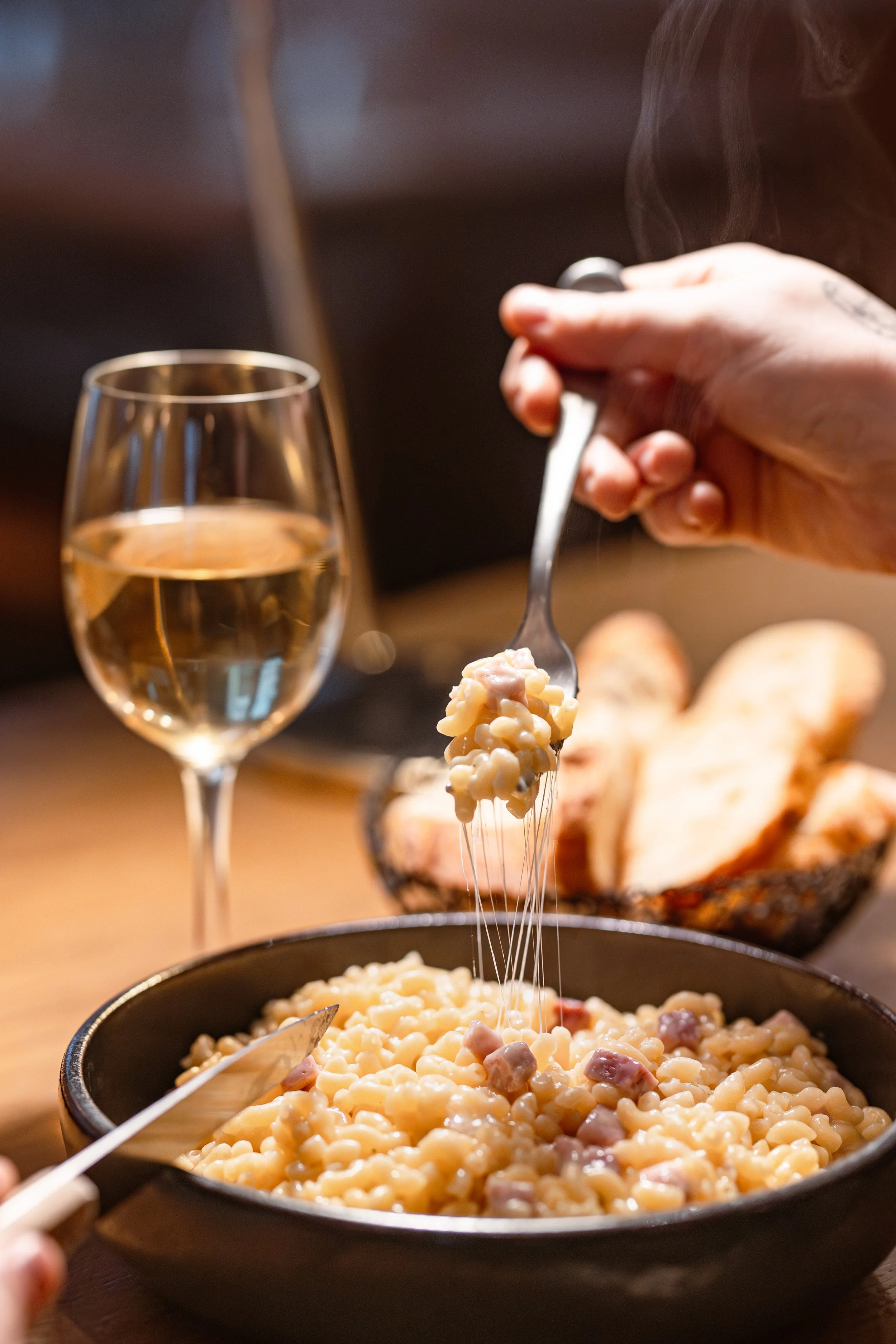 Une main tient une fourchette avec des pâtes au fromage et au jambon dans un bol noir. Sur la table, une verre de vin blanc et du pain en arrière-plan.