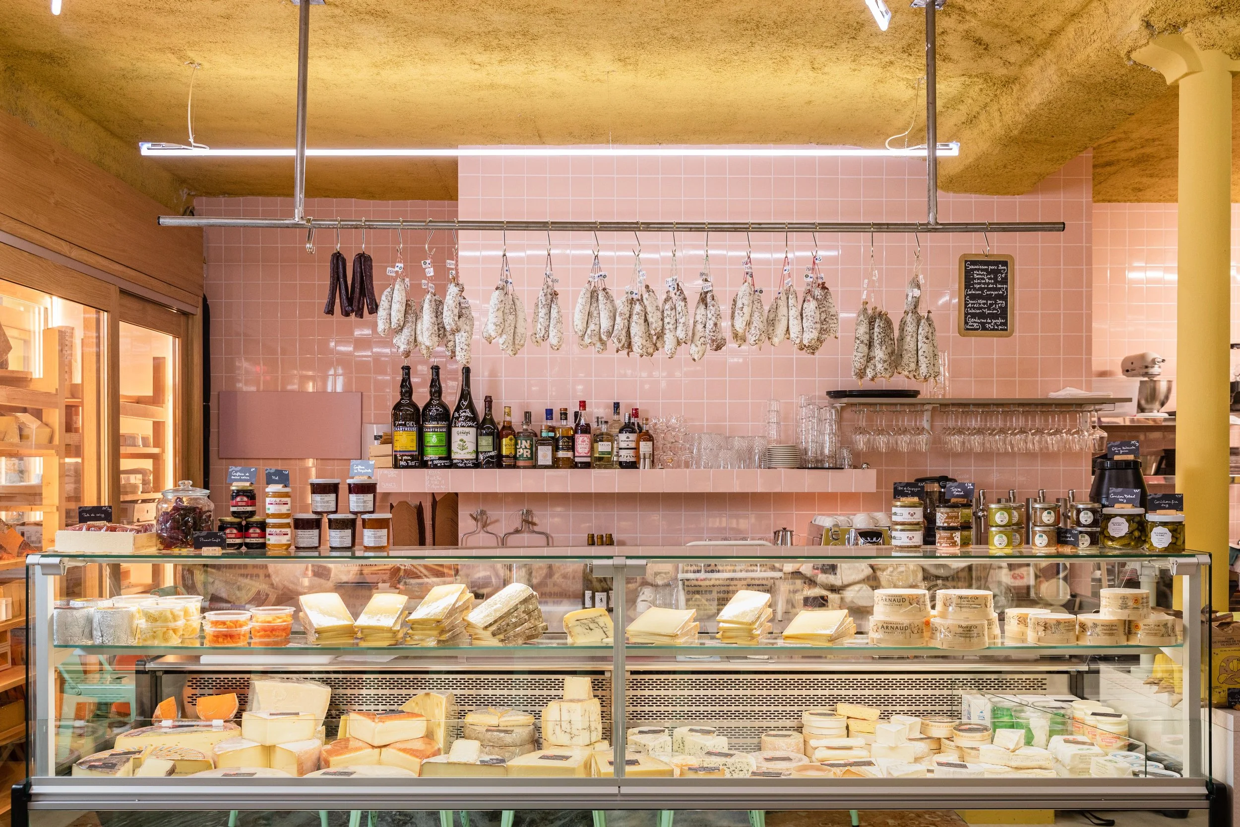 Vitrine de la fromagerie Monbleu Père lacheese rempli de fromage avec des saucissons suspendu en hauteur, le mur est rose dans le fond et le plafond est jaune
