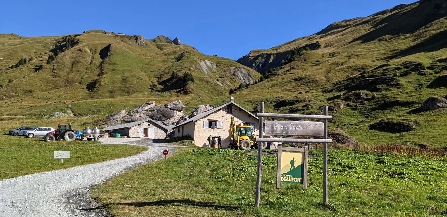 Paysage avec de grandes collines vertes, un chemin qui mène a plusieurs maison, il y a un tracteur et des voitures, le ciel est bleu et il fait très beau