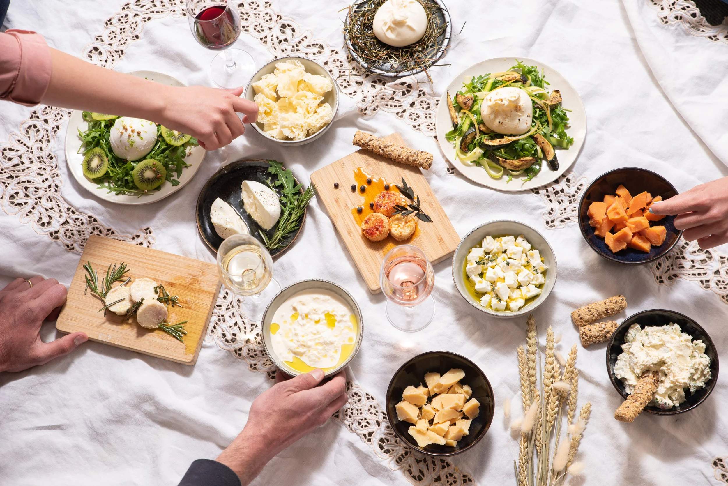 Un repas convivial sur une table habillée d'une nappe blanche avec un motif en dentelle, comprenant diverses assiettes de fromages, un plat de légumes, un œuf poché, un verre de vin rouge, et des mains qui se servent.