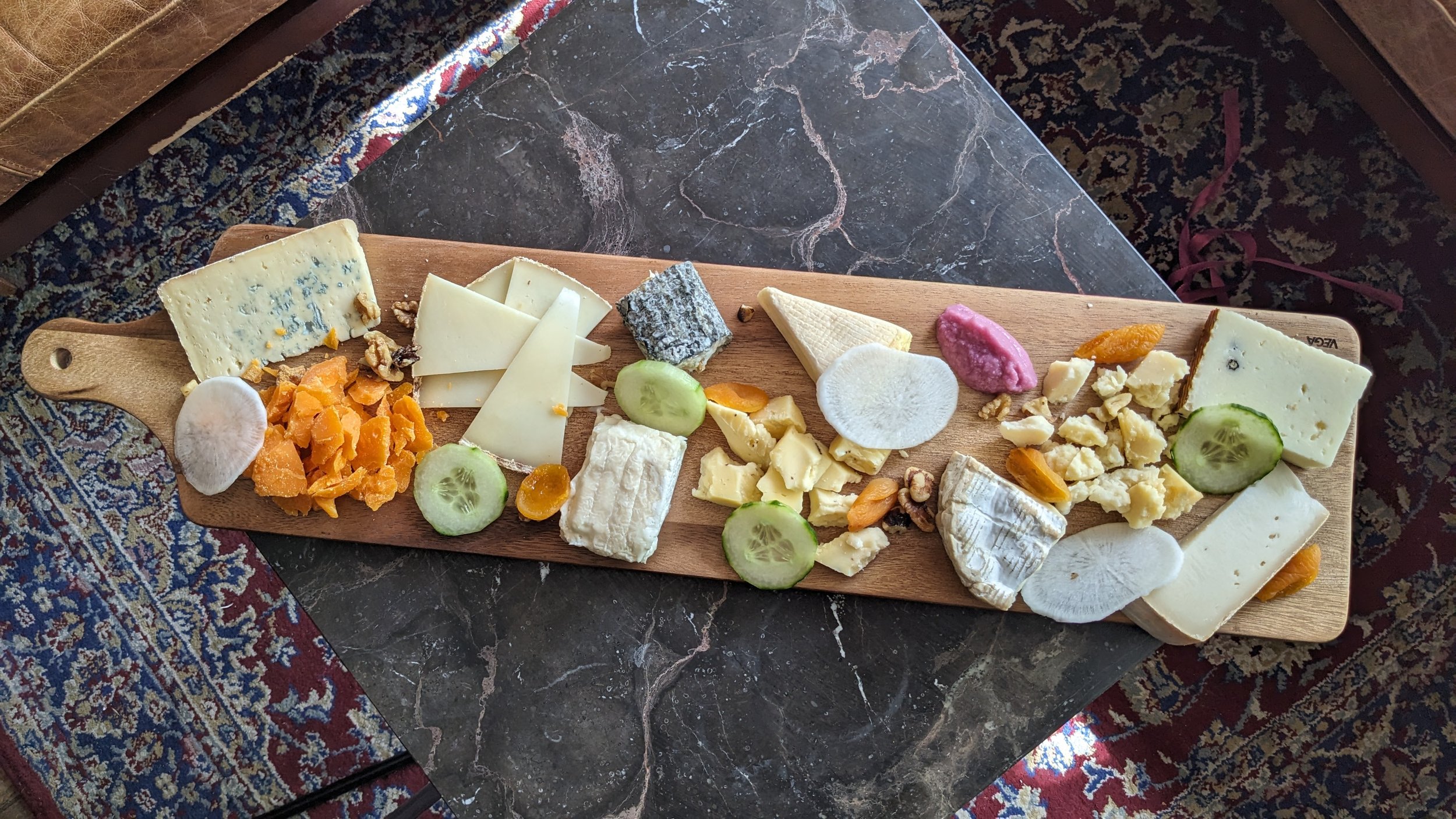 Une grande planche en bois avec un assortiments de plusieurs fromage, avec des concombres, sur une table en marbre gris foncé en arrière plan il y a un tapis
