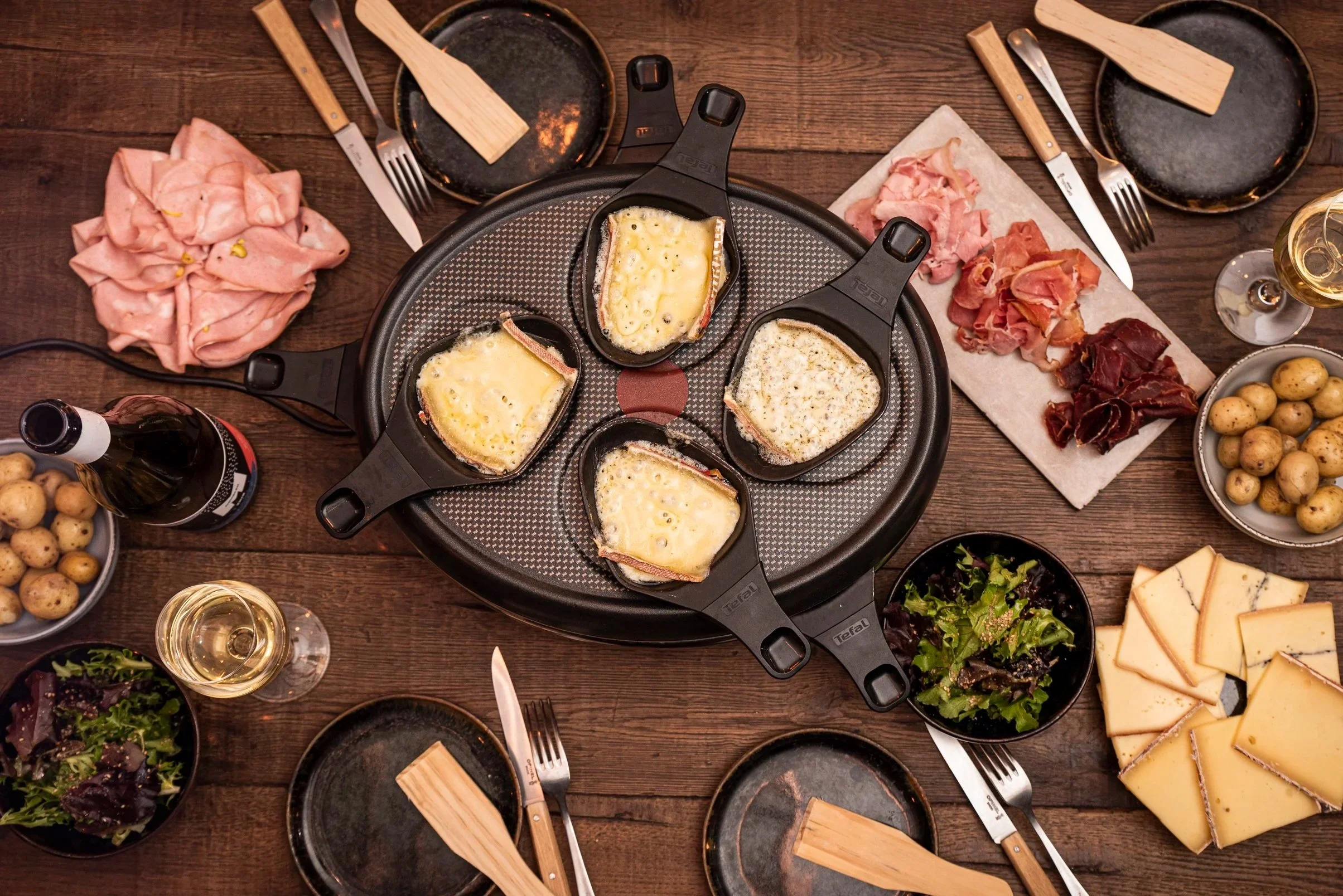 Table dressée pour un repas avec un appareil de Raclette, des assiettes, des charcuteries, des fromages, des pommes de terre et des boissons