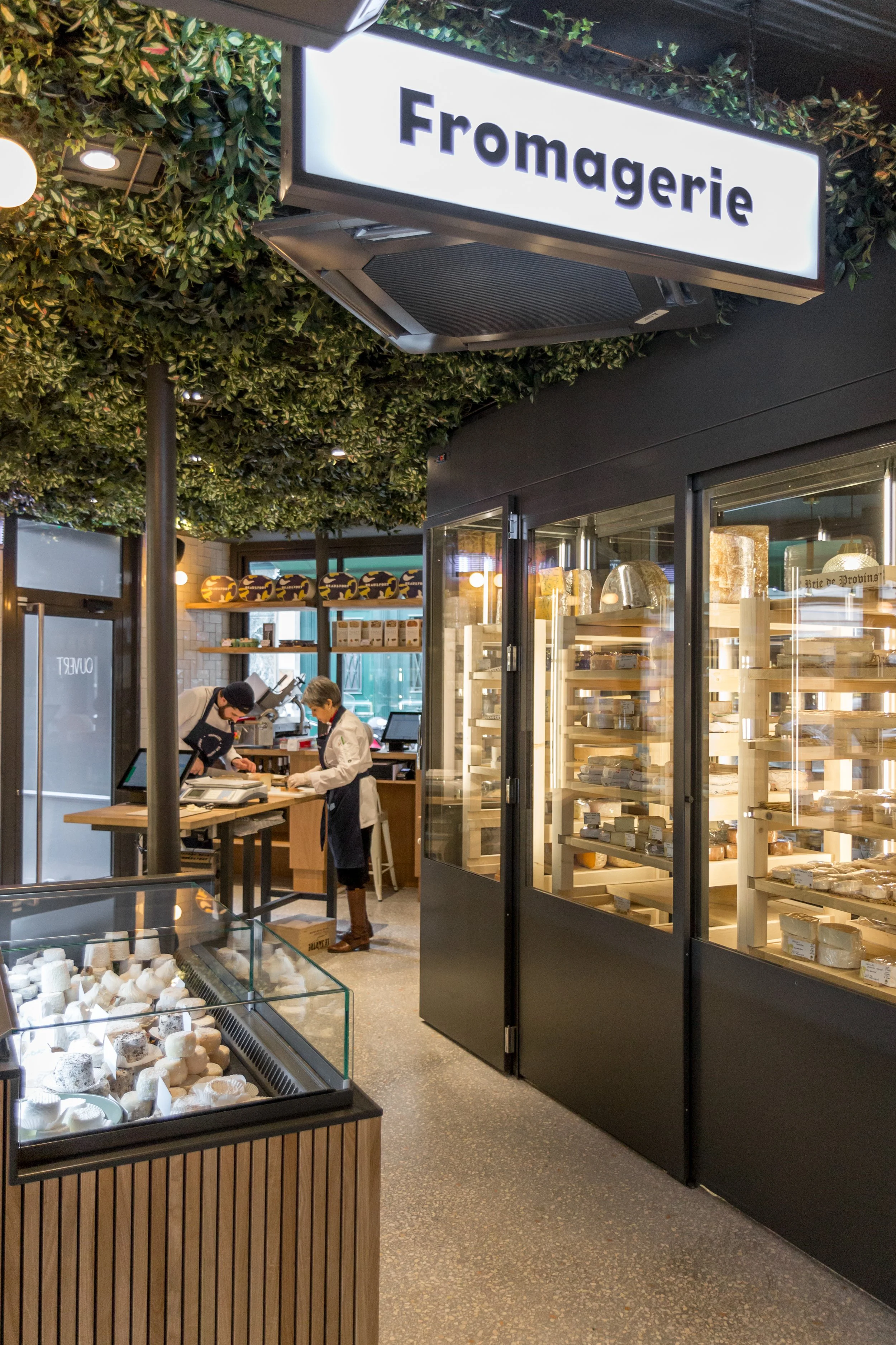 Une fromagerie élégante avec un panneau lumineux indiquant 'Fromagerie' et un intérieur bien éclairé où des employés travaillent derrière un comptoir de fromage, de nombreux fromages exposé en vitrine.