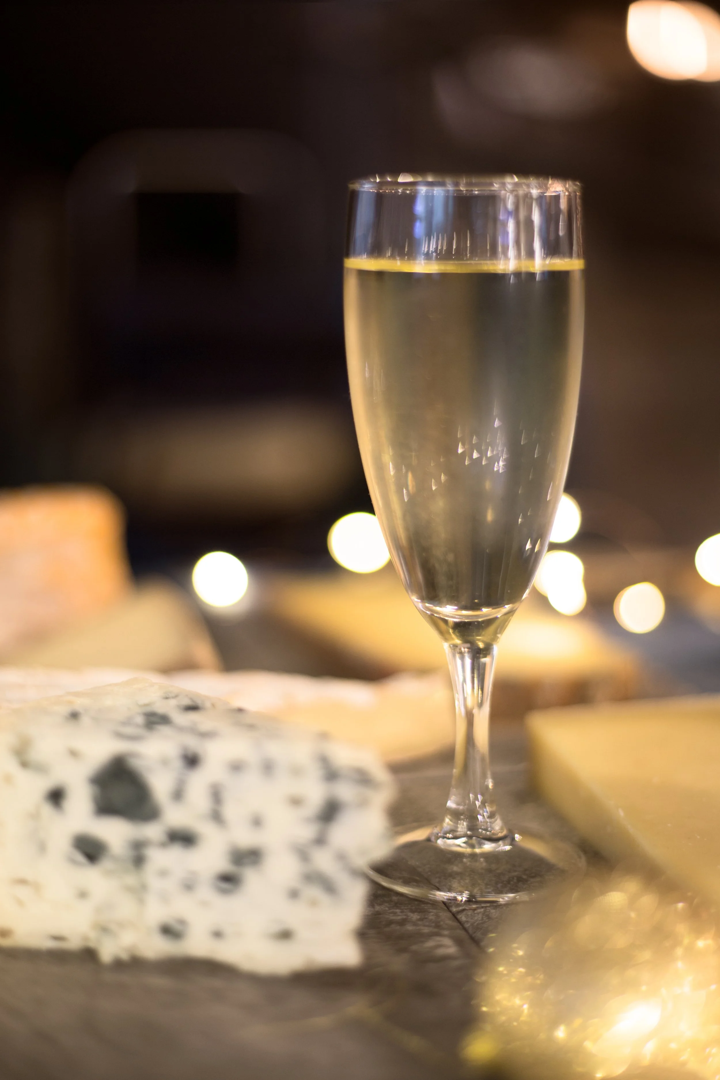 Verre de champagne placé sur une surface en pierre avec du fromage en arrière-plan, ambiance chaleureuse et lumineuse.