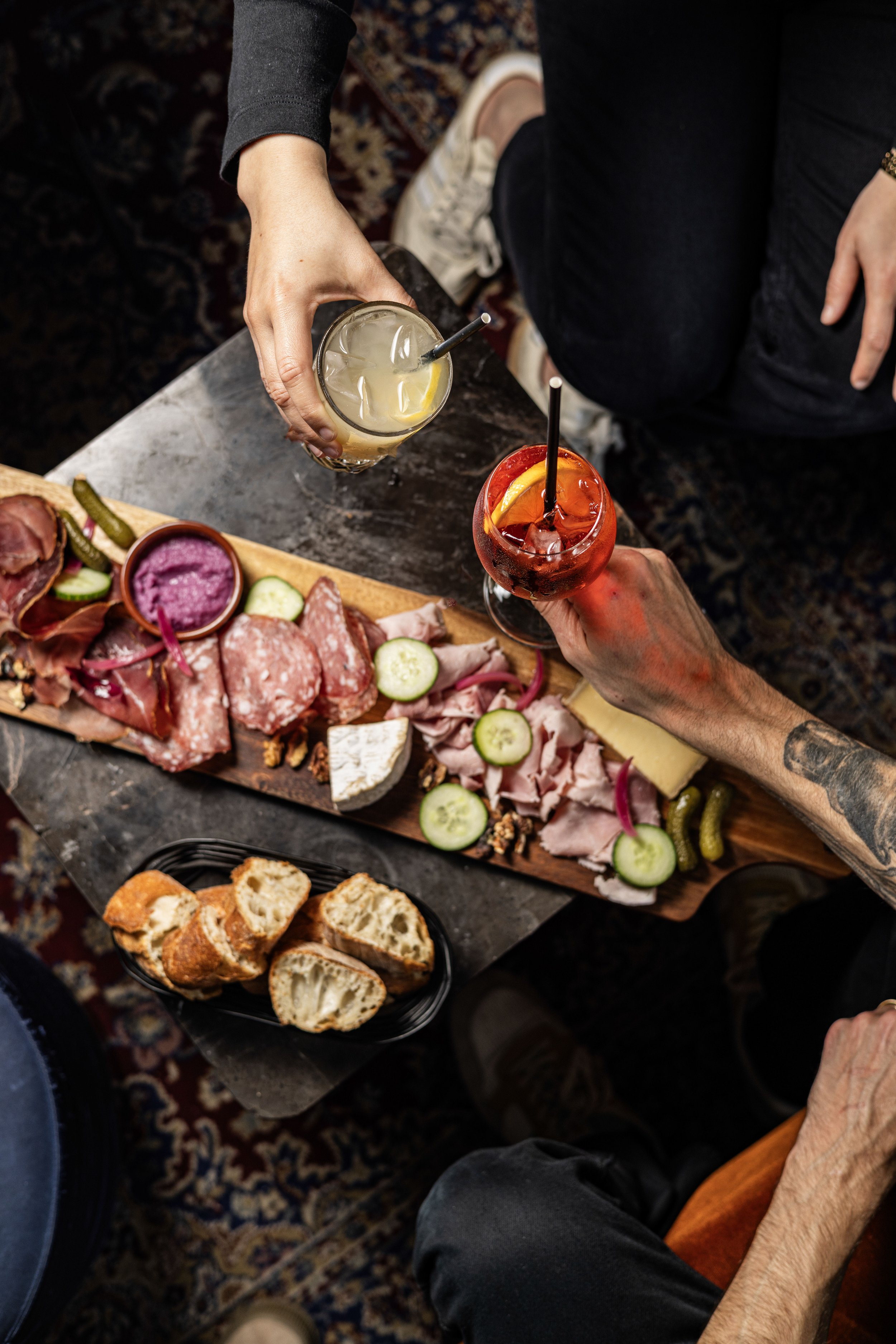 Un apéritif entre deux personne minimum, elles tiennents des cocktails dans leur main, avec une planche mixte, charcuterie et fromage et une corbeille de pain