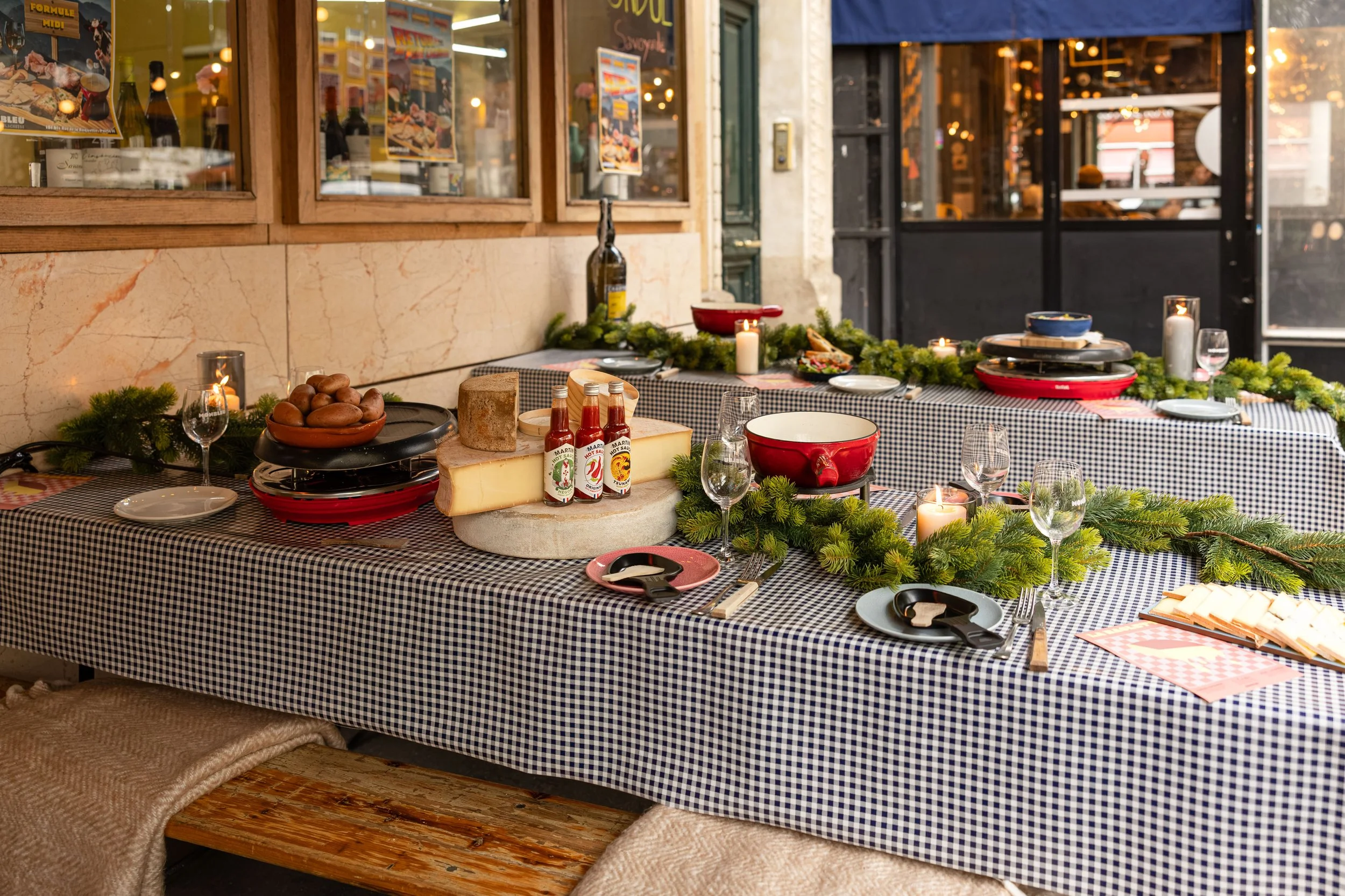Terrasse du restaurant MONBLEU Père Lacheese à Paris 11e, avec tables dressées pour une raclette et une fondue, fromages, appareils à raclette et verres à vin.