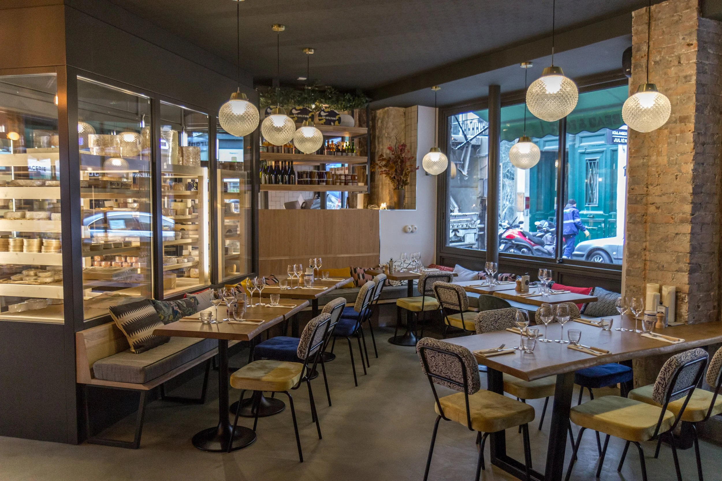 intérieur d'un restaurant chaleureux, avec une banquette et des coussins et des chaises en face, les tables sont dressées, on voit deux grandes vitrines rempli de fromages sur la partie gauche de l'image ainsi que du vin et des accompagnements.