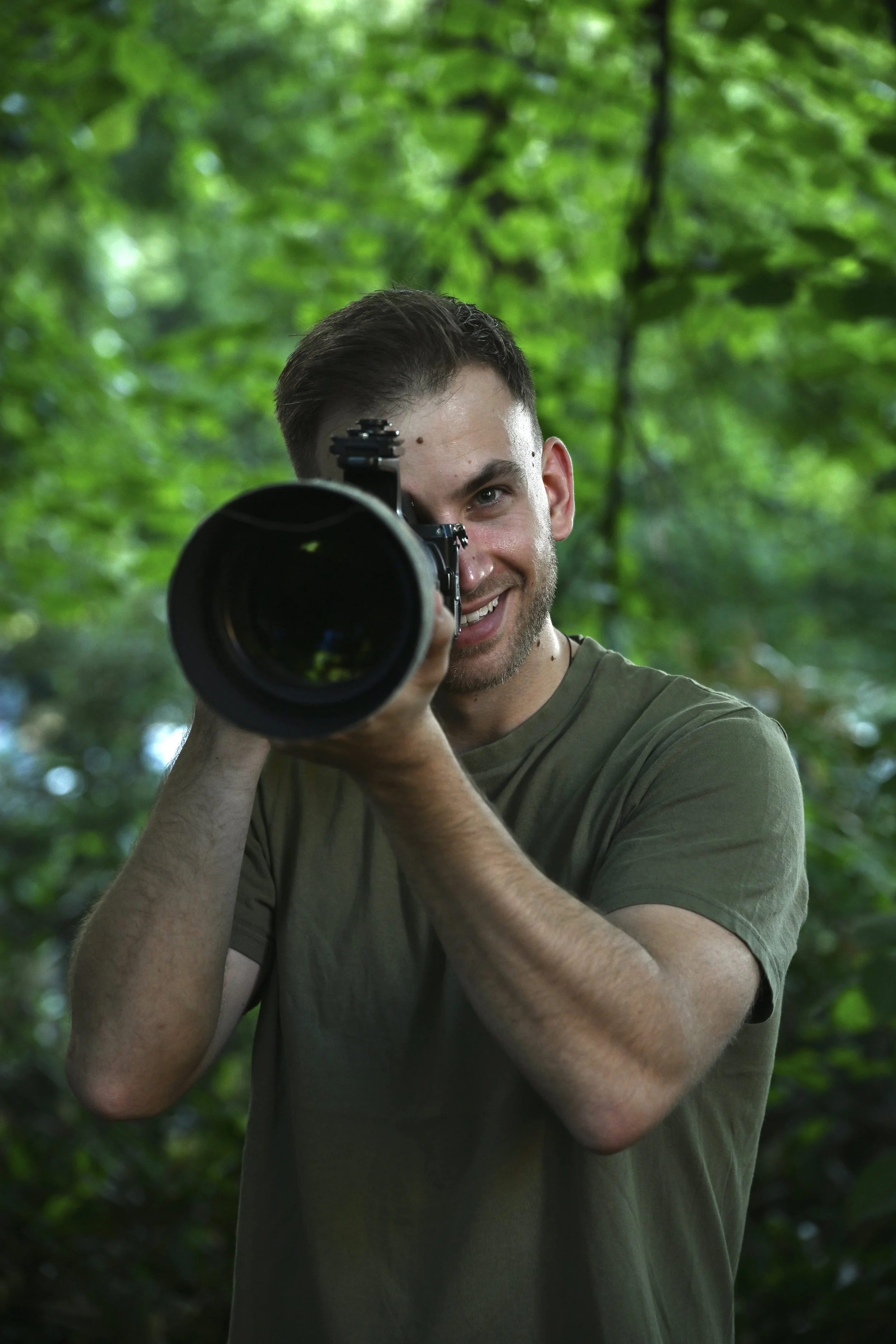 A man with short brown hair and a slight beard, wearing a green t-shirt, is smiling while holding a camera with a large telephoto lens, pointing it towards the viewer, with a blurred green forest background.