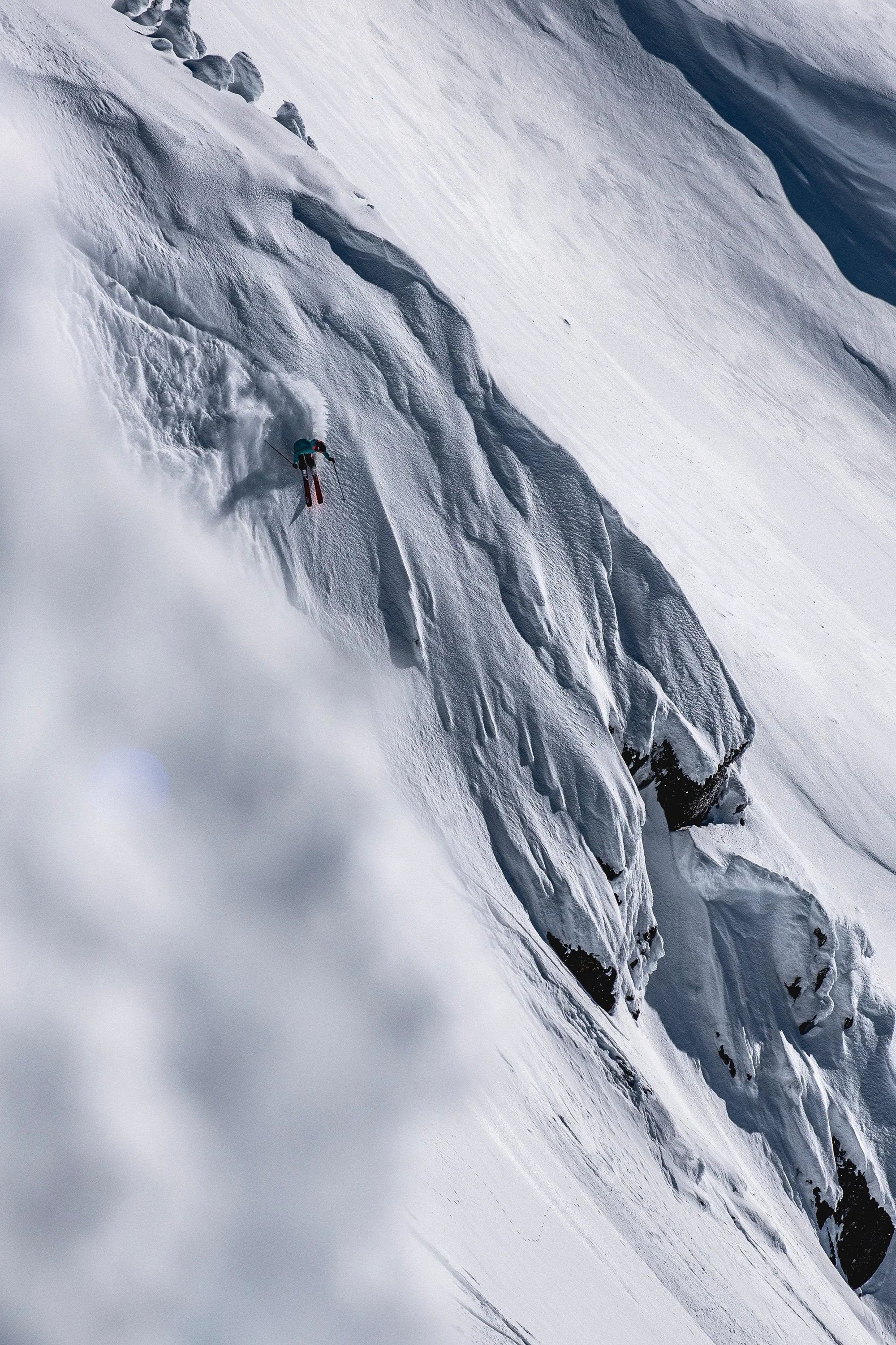 A skier, Kayley Sherlock, in a teal jacket and black pants, skiing down a steep snow-covered mountain with rugged cliffs and snowdrifts.