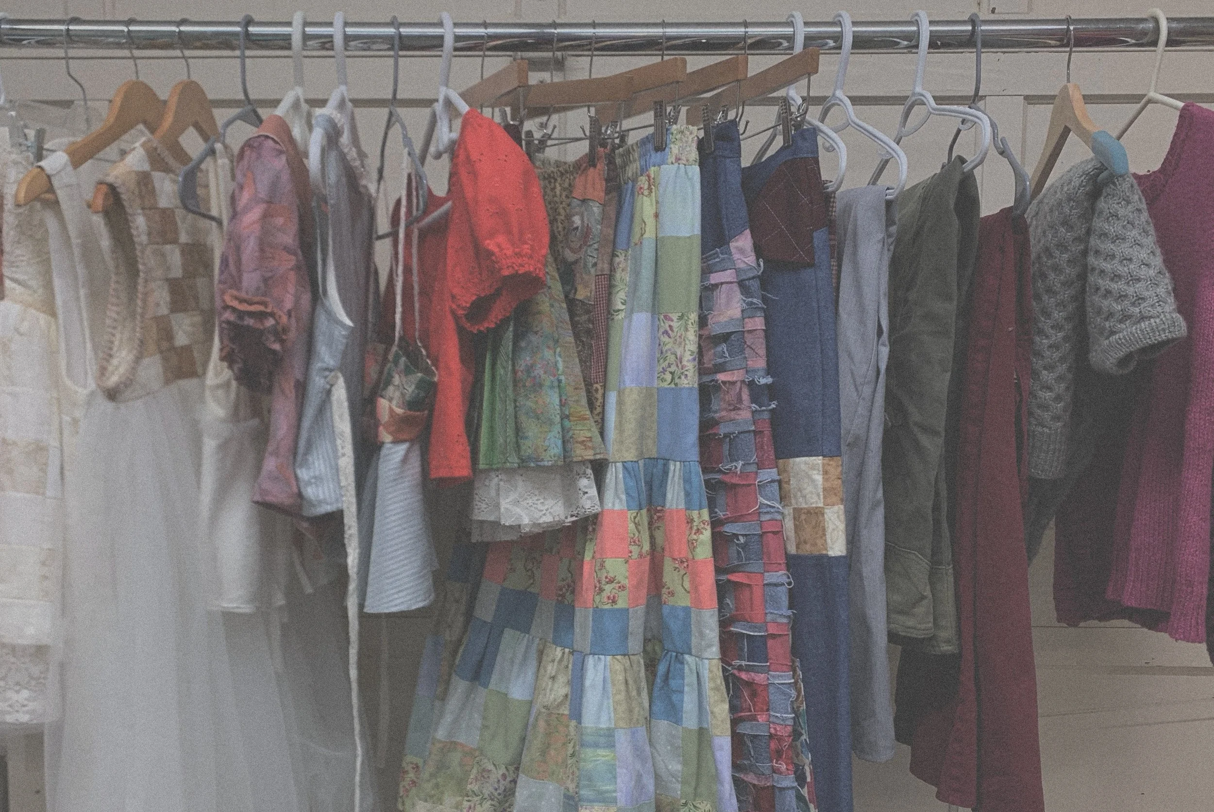 Clothing items hanging on a metal rod in a closet, including dresses, pants, skirts, and sweaters in various colors and patterns.