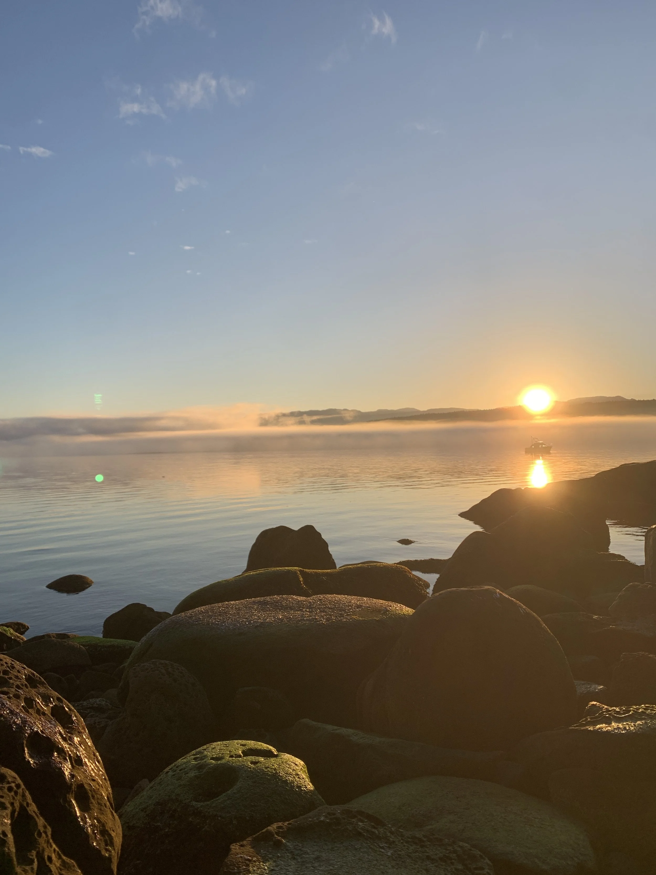 Sunset over a calm ocean with rocks in the foreground and a boat on the water.