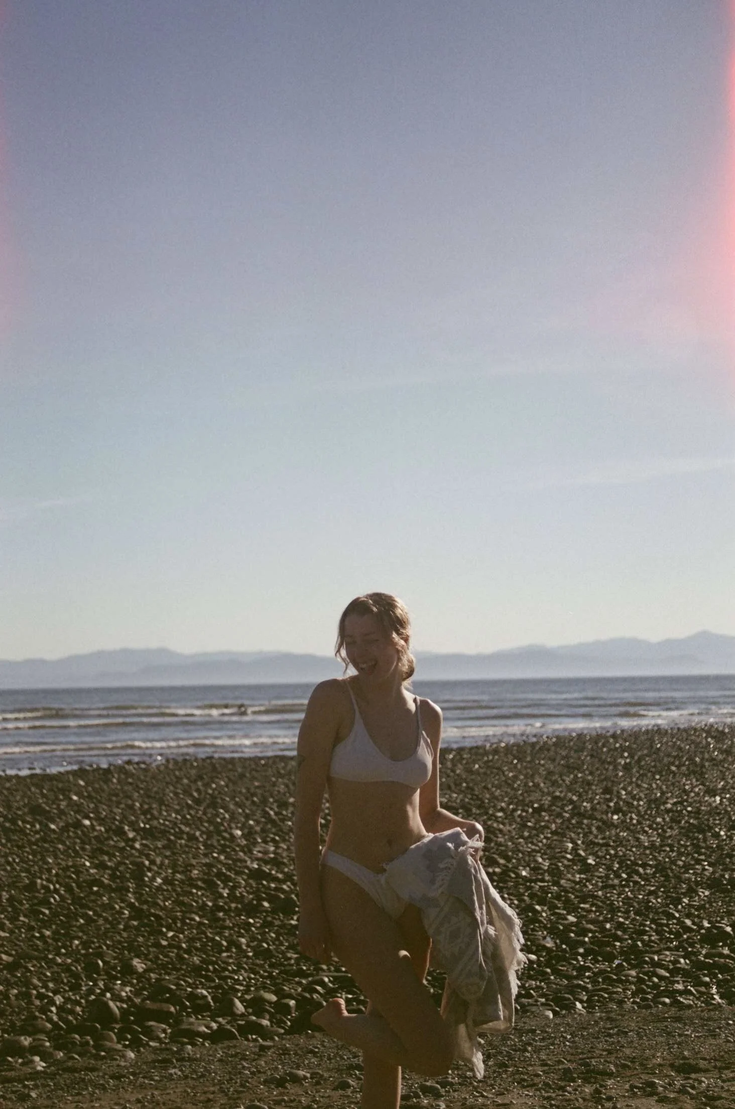 A film photo of a woman in a white bikini standing on a pebble beach, smiling, holding a towel, with the ocean and mountains in the background during sunset or sunrise.