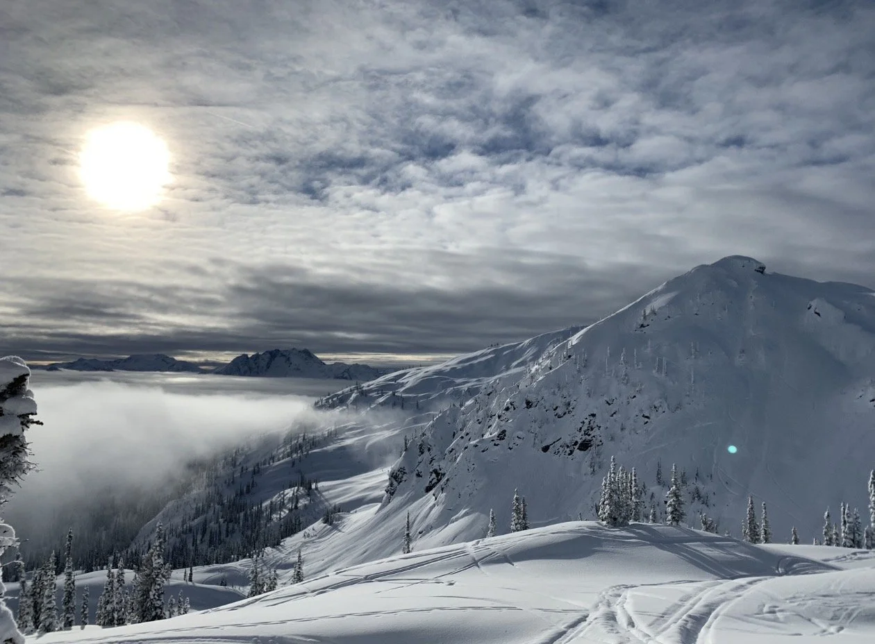 Snow-covered mountain landscape with ski trails, evergreen trees, and a cloudy sky with the sun shining through.