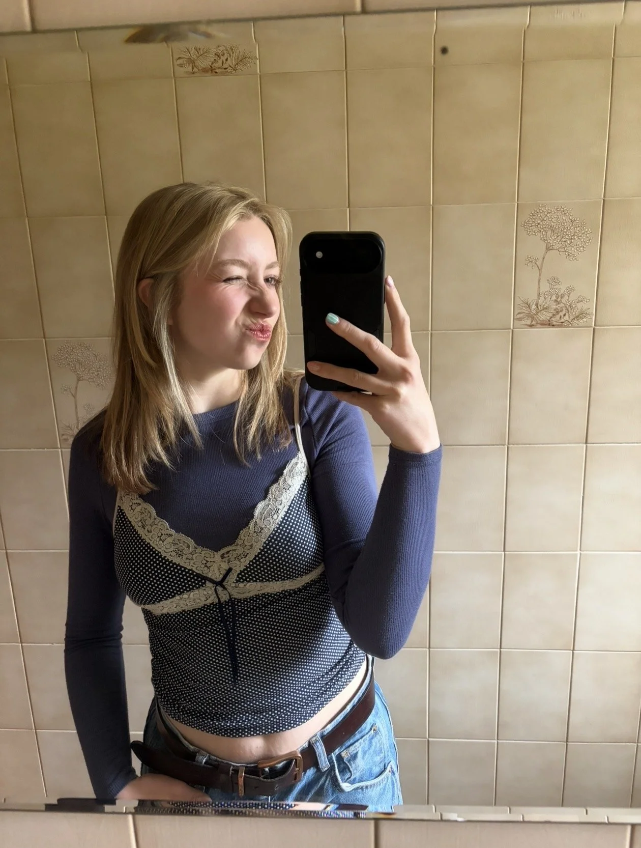 Young woman taking a mirror selfie in a bathroom, wearing a long sleeve navy blue top with lace and polka dot details, and blue jeans.