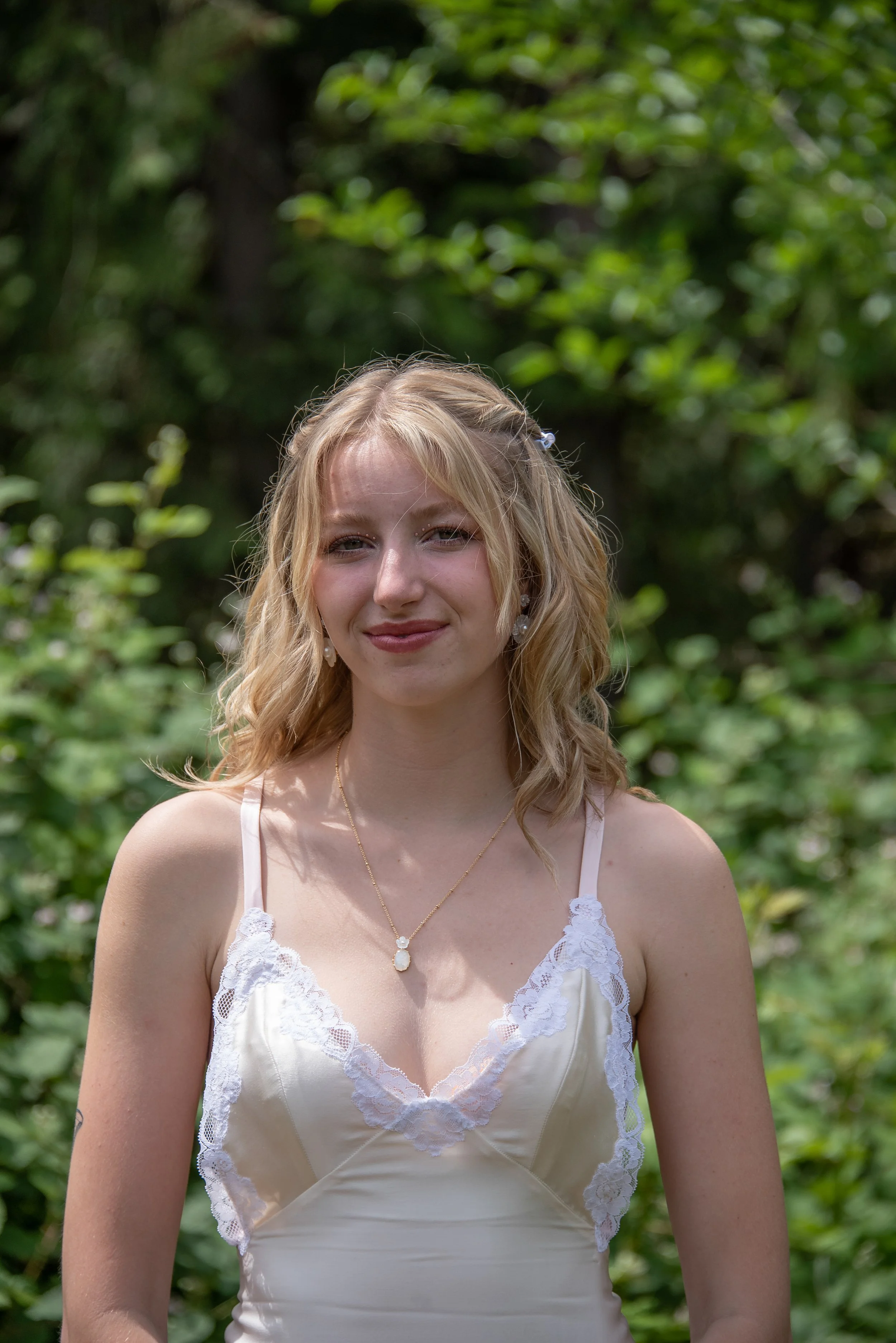 A young woman with blonde hair styled in loose waves, wearing a white satin dress with lace detailing at the bust and spaghetti straps, standing outdoors in front of a green leafy background.