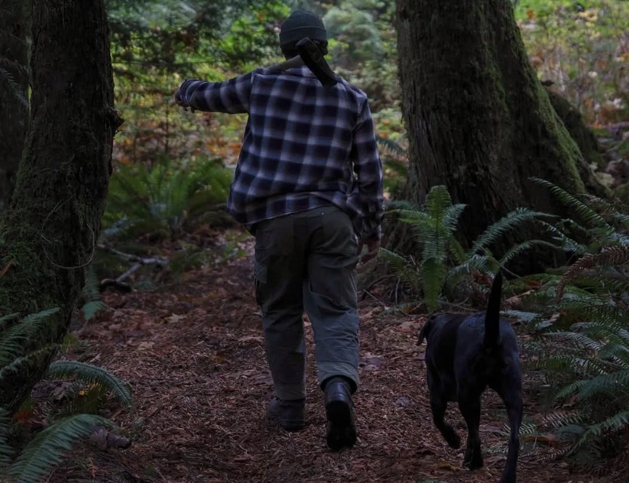 A person wearing Lupine Studios pants walking a dog on a forest trail surrounded by trees and foliage. 