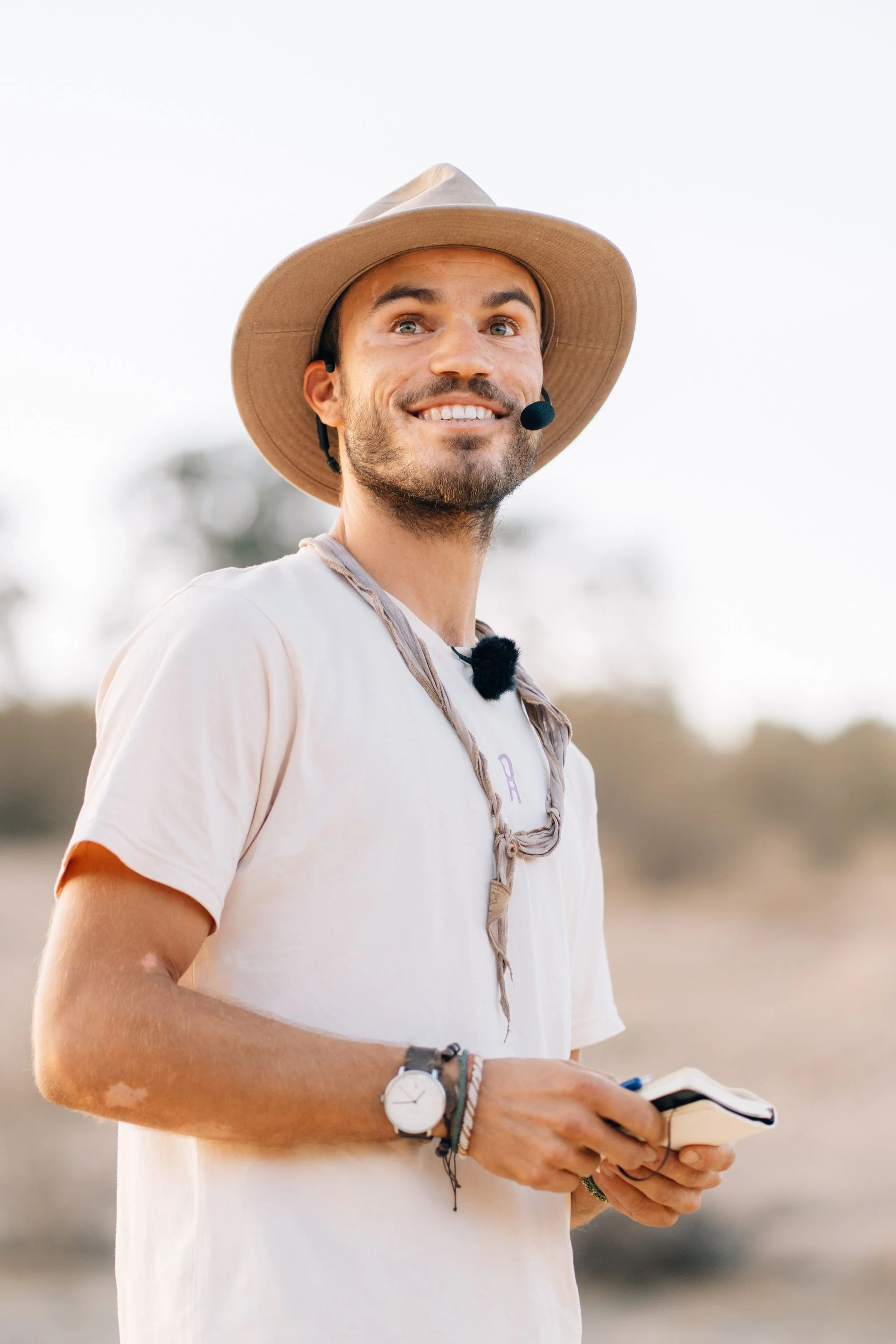 Smiling man wearing a wide-brimmed tan hat and a beige t-shirt with a headset microphone, holding a notebook, outdoors during daytime.