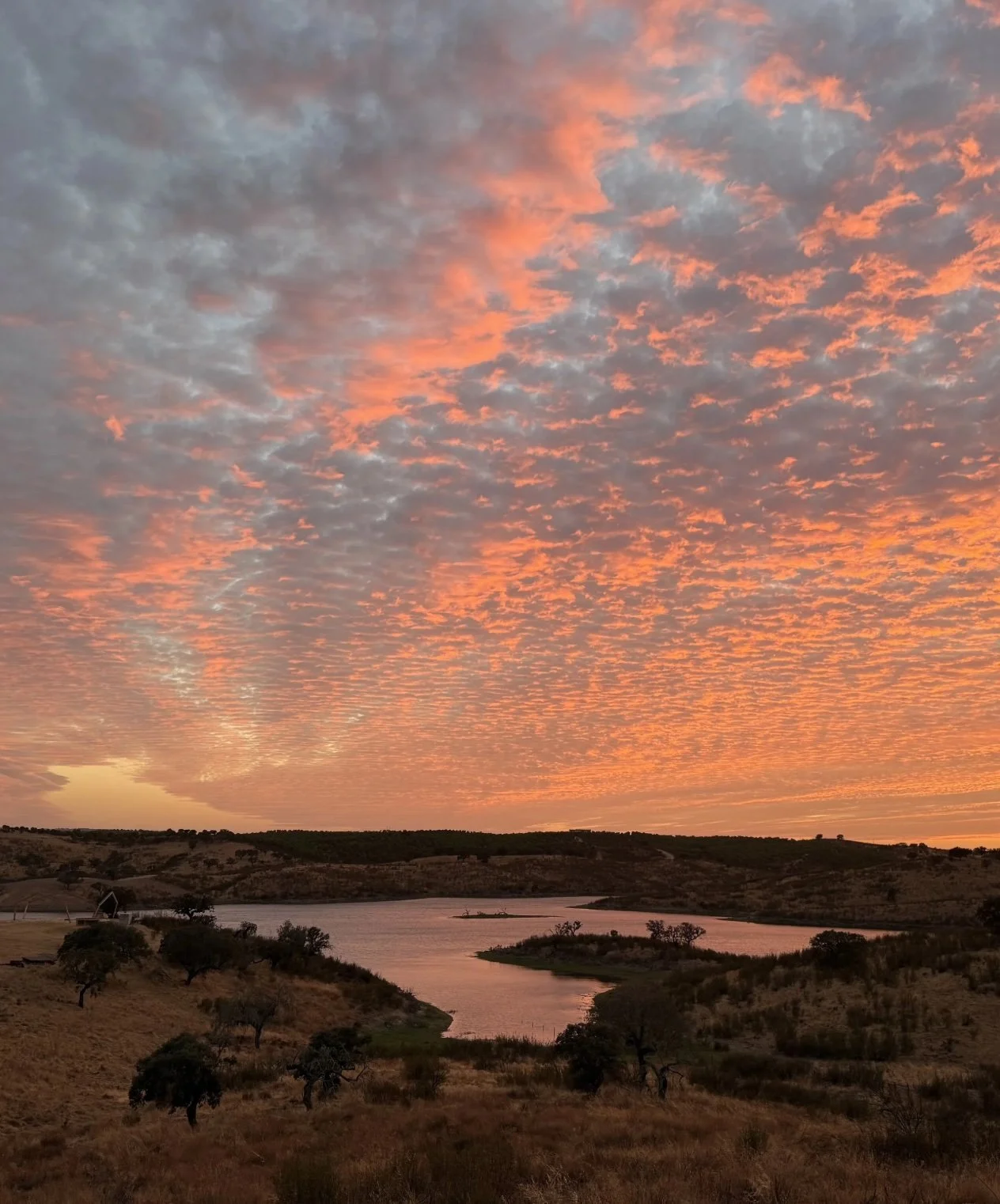 A landscape scene at sunset with a river flowing through dry, hilly terrain and a sky filled with orange and gray clouds.