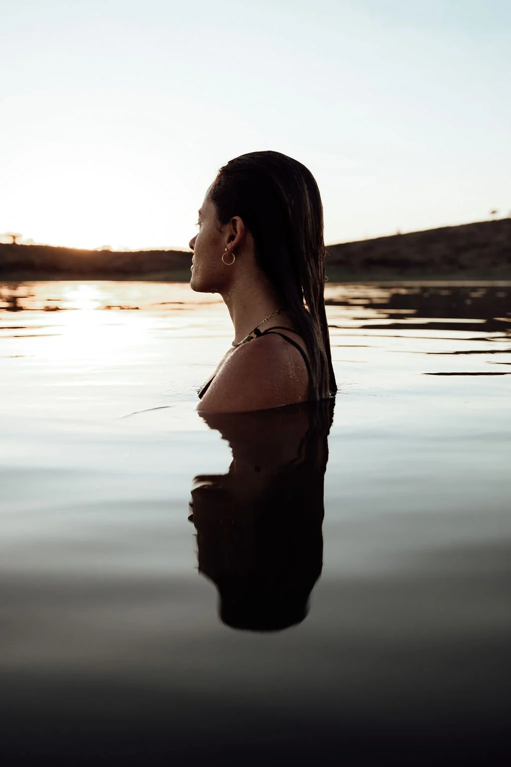 A person with dark, wet hair and earrings is submerged up to their shoulders in a calm body of water during sunset, with the person's profile visible against the bright sky.