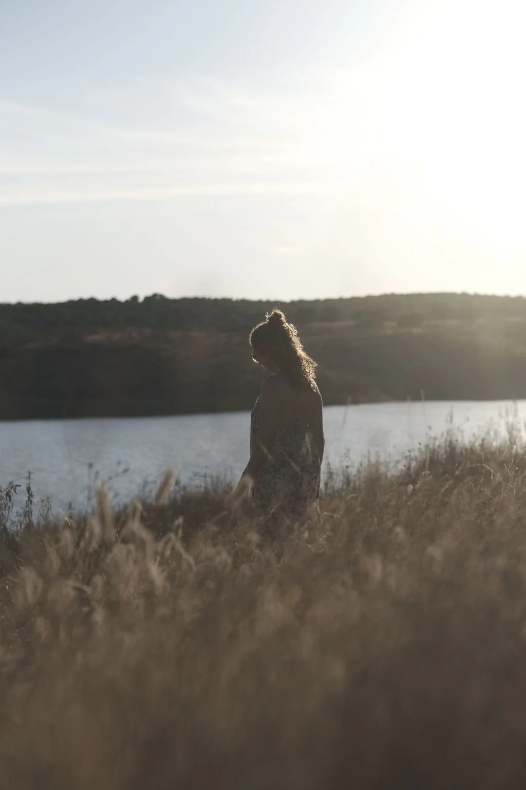 A woman standing outdoors near a body of water during sunset or sunrise, with tall grass in the foreground and a hill in the background.