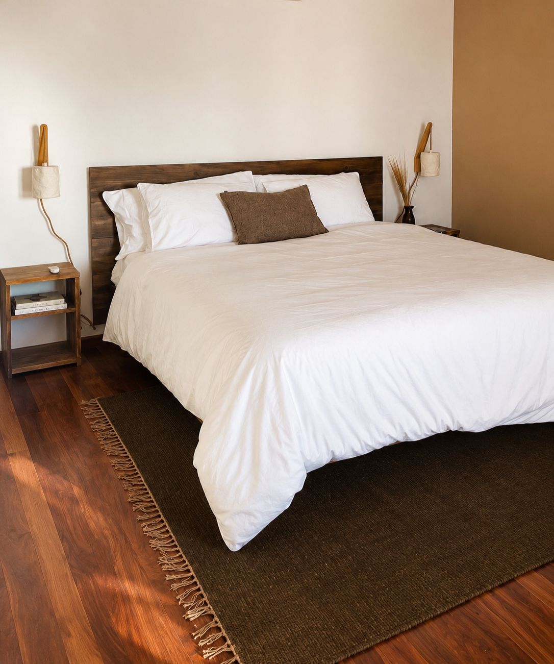 A neatly made bed with white bedding, a brown throw pillow, and dark brown wooden headboard, flanked by side tables with wall-mounted lamps in a minimalist bedroom with hardwood floors and beige walls.