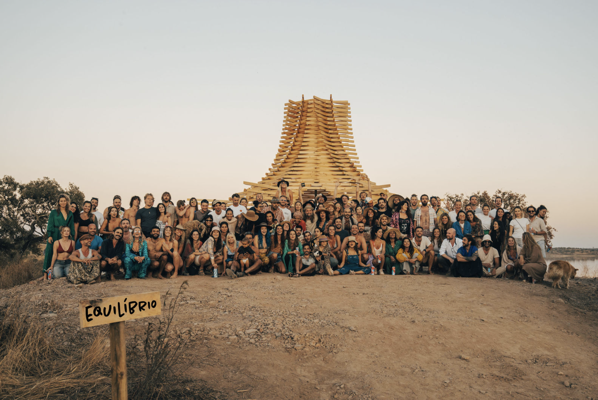 A large group of people gathered outdoors in front of a wooden art installation resembling a large, curved architectural structure. There is a wooden sign in the foreground that reads 'EQUILIBRIO'. The background features trees and a clear sky.