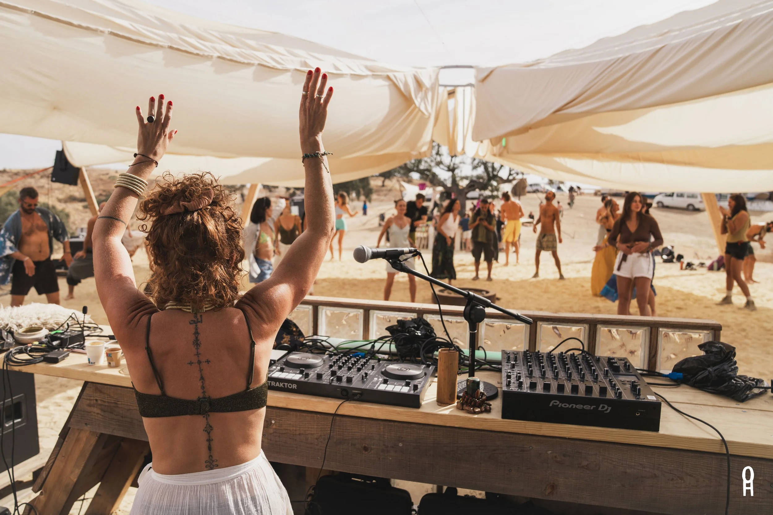 A woman with curly hair and tattoos raised her hands in front of a DJ setup at an outdoor festival or beach party, with people dancing and socializing in the background under large white fabric canopies.