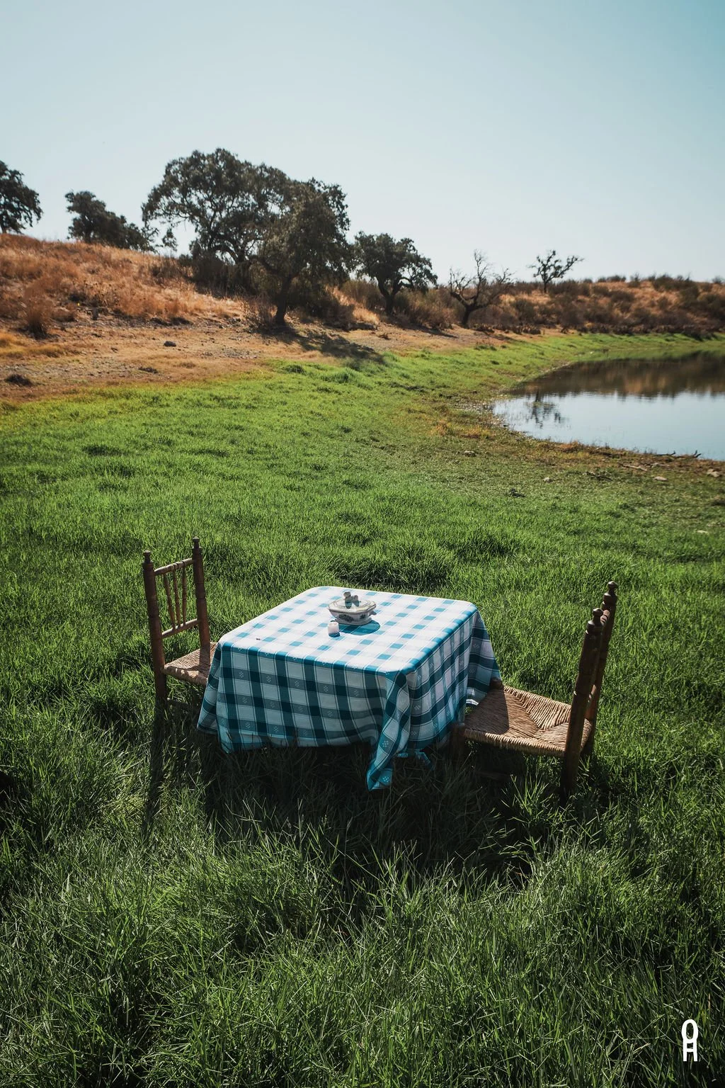 A table with a blue checkered tablecloth and two wooden chairs set outdoors near a small lake, with a rocky and grassy landscape in the background under a clear sky.