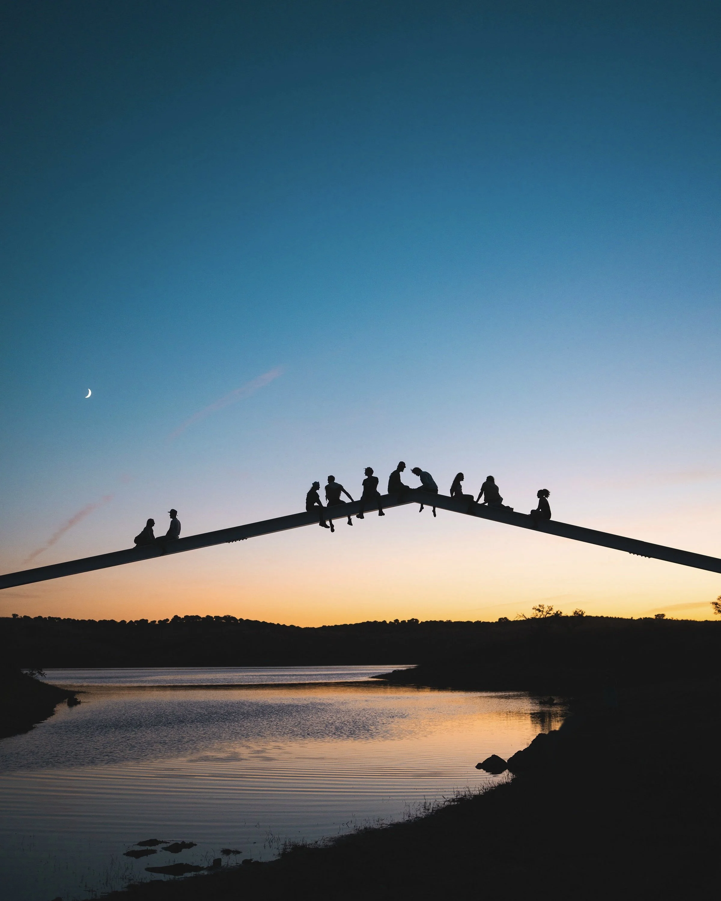 Silhouette of people sitting on a bridge at sunset over a river, with a crescent moon in the sky.