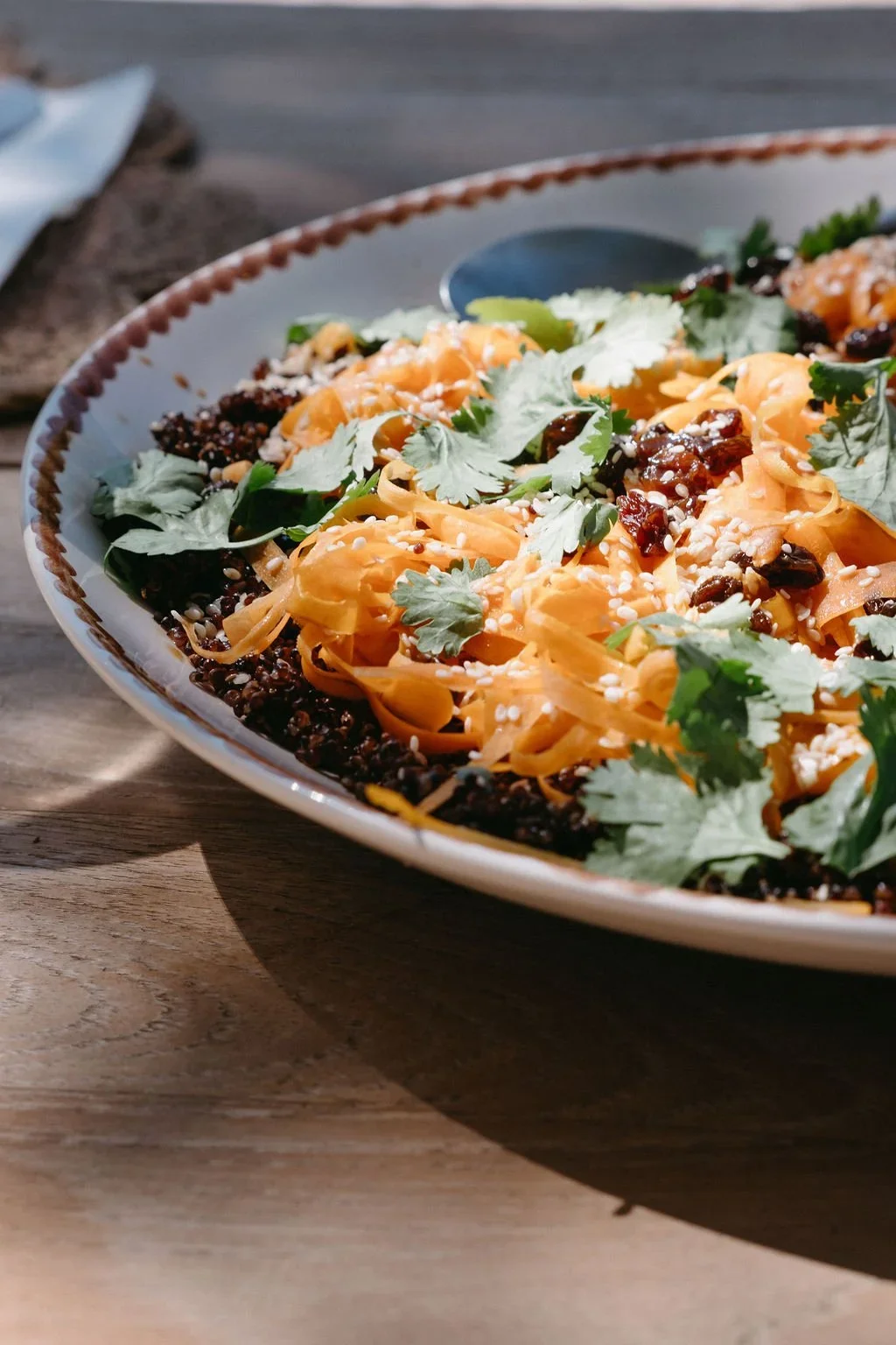A close-up of a bowl of pasta topped with shredded cheese, black beans, chopped herbs, and sesame seeds, placed on a wooden table with a spoon nearby.