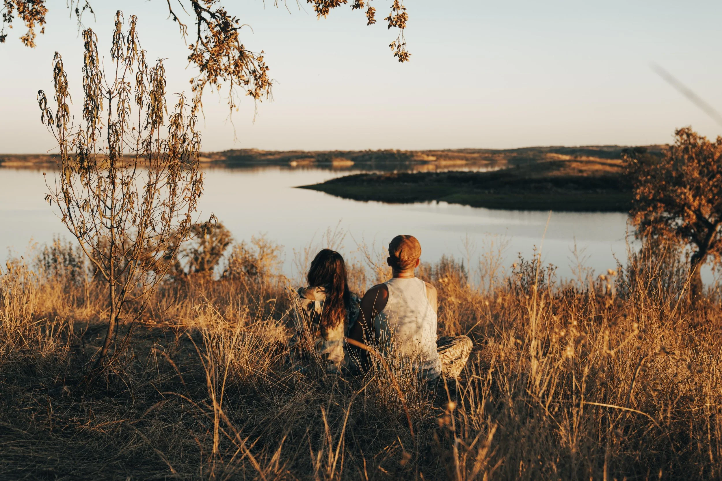 Two people sitting on a grassy hillside overlooking a lake during sunset, surrounded by trees with autumn leaves.