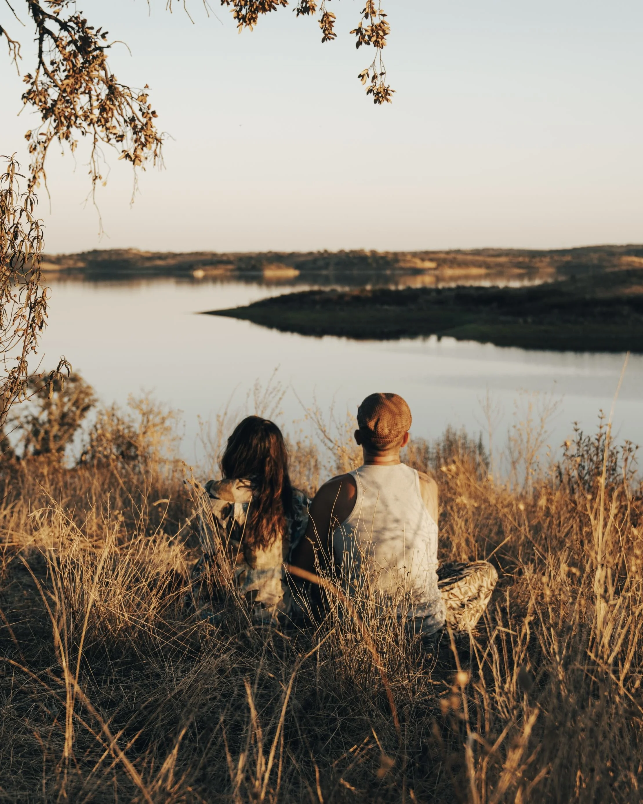 Two people sitting in tall grass near a body of water, watching the sunset with trees in the foreground and background.