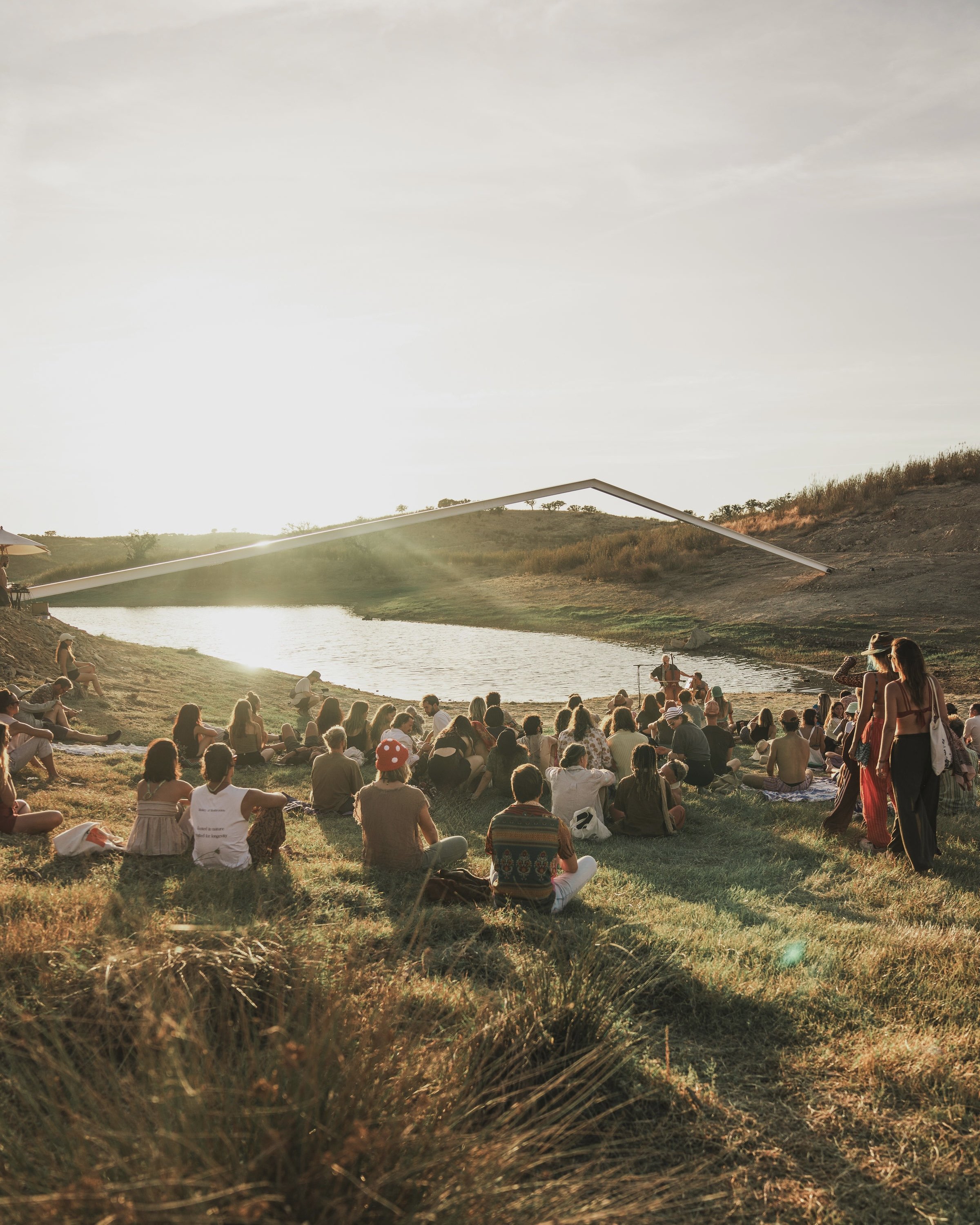 People gathered outdoors near a lake during sunset, with some sitting on the grass and others standing, and a person speaking into a microphone near the water.