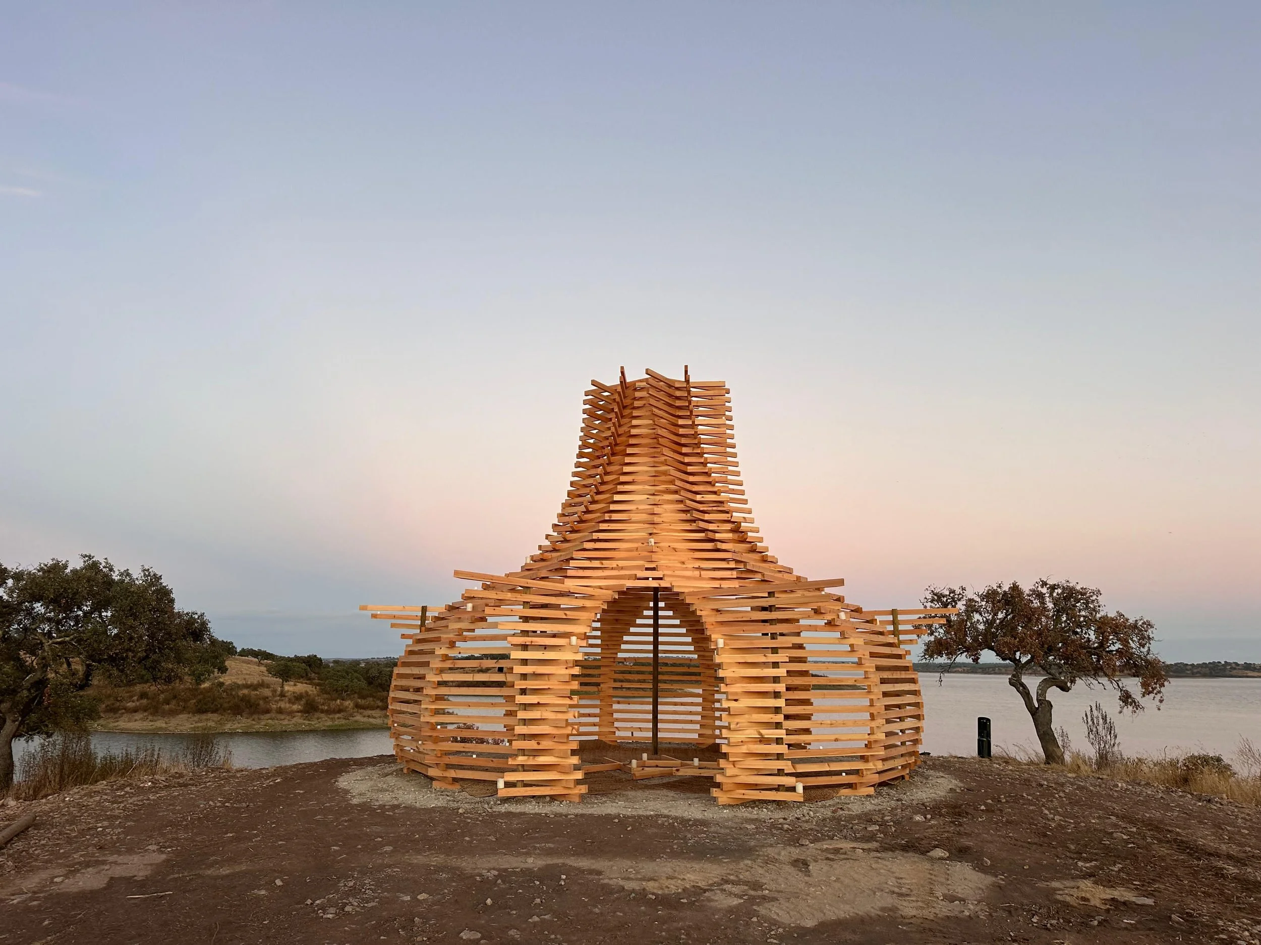 A wooden sculpture resembling a tower or chimney near a body of water, with trees and a sunset sky in the background.