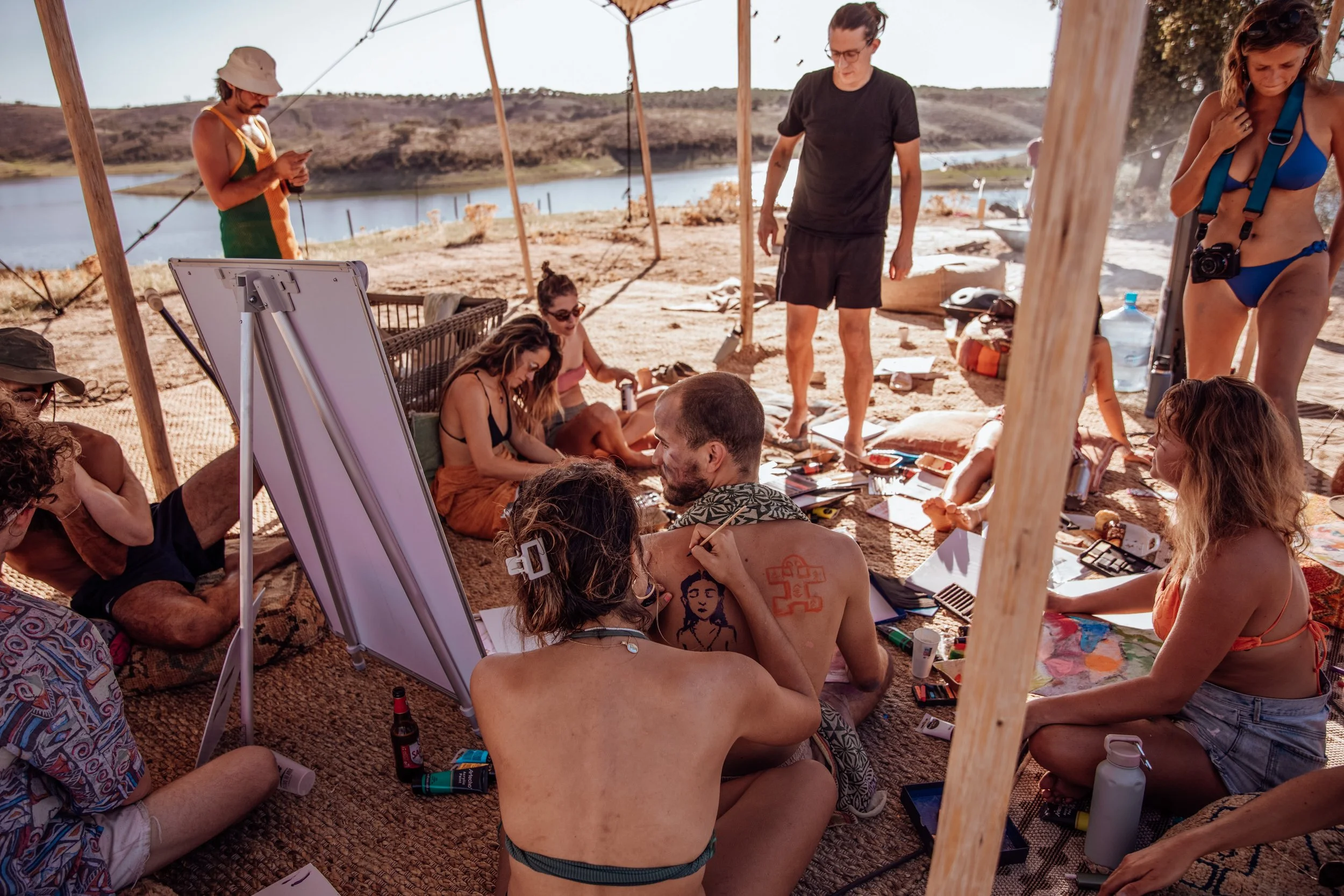 A group of people gathered under a canopy outdoors by a river, sitting on the ground, drawing and engaging in various activities.