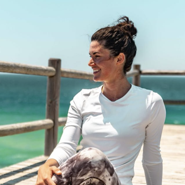 Woman sitting on a wooden pier by the ocean, smiling and looking to her right, with her hair in a bun and wearing a white long-sleeve shirt.