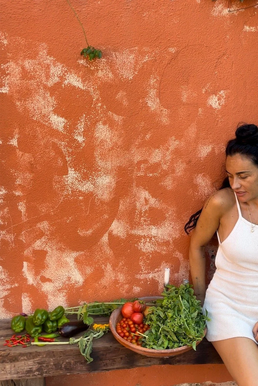 Woman in a white dress sitting against an orange wall beside a rustic wooden table with fresh vegetables and herbs, including bell peppers, eggplant, tomatoes, and chili peppers.