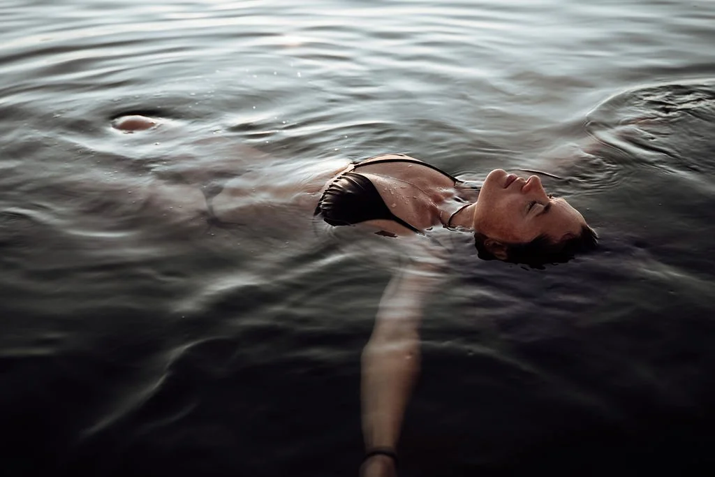A woman floating on her back in dark water with eyes closed, relaxed, wearing a black top.