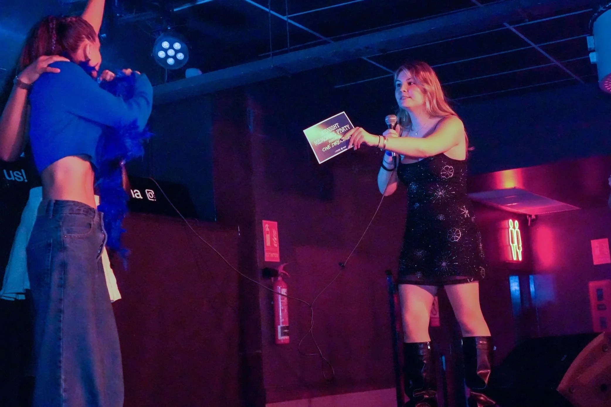 A woman in a black dress holding a microphone and a card, standing on a stage with colorful lights and a dark background. Another person, partially visible, is facing her.