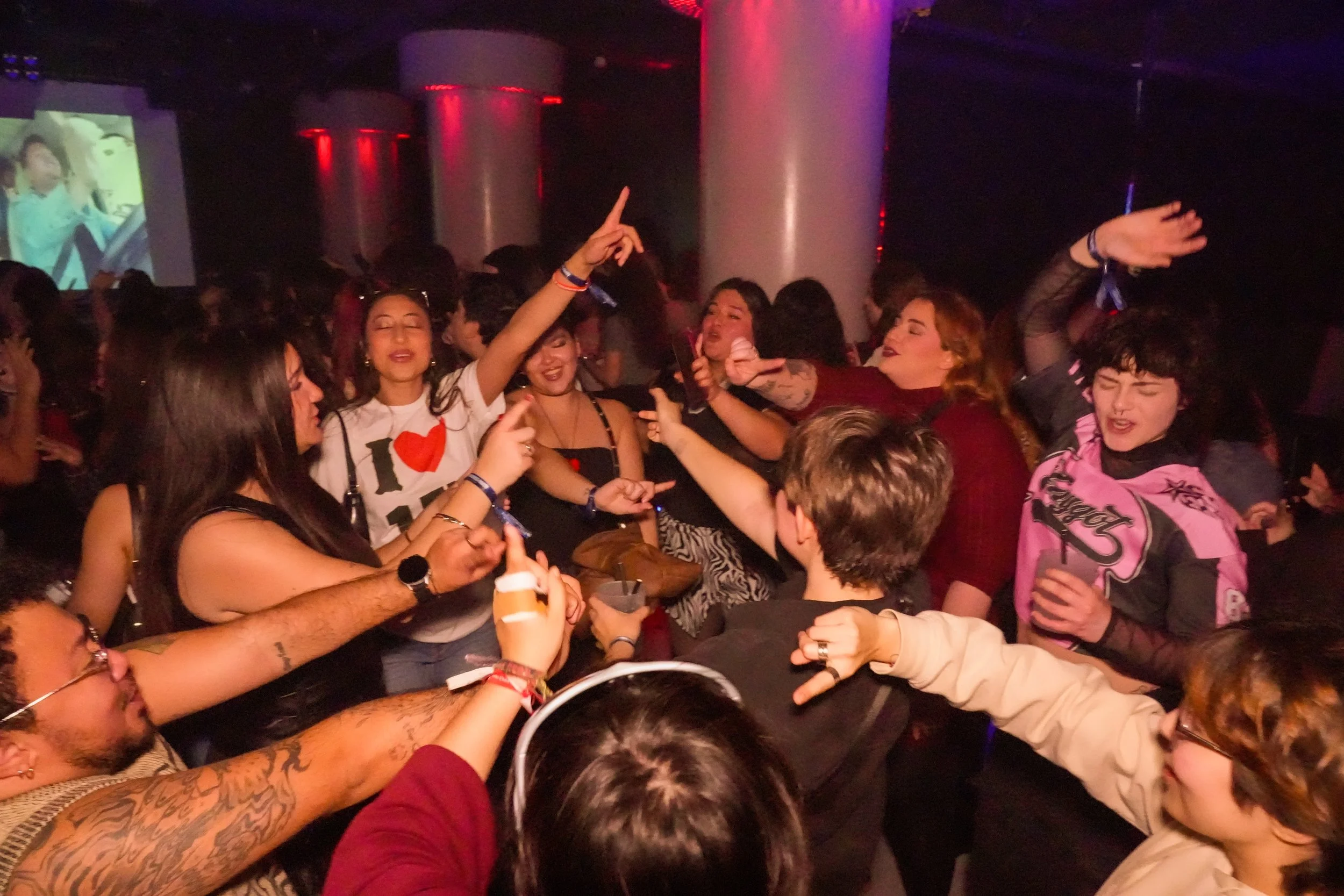 Crowd of young people at a lively party or nightclub, dancing and socializing, with colorful dim lighting and a large screen in the background.